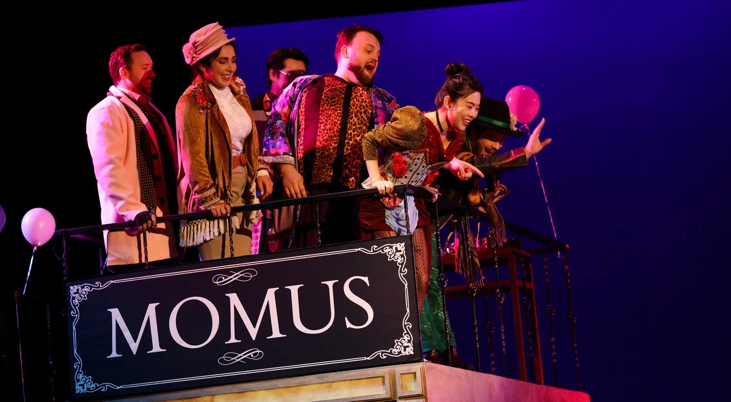 A group of actors on stage dressed in colorful, vintage costumes, leaning on a balcony railing with a "MOMUS" sign, celebrating a theatrical performance.