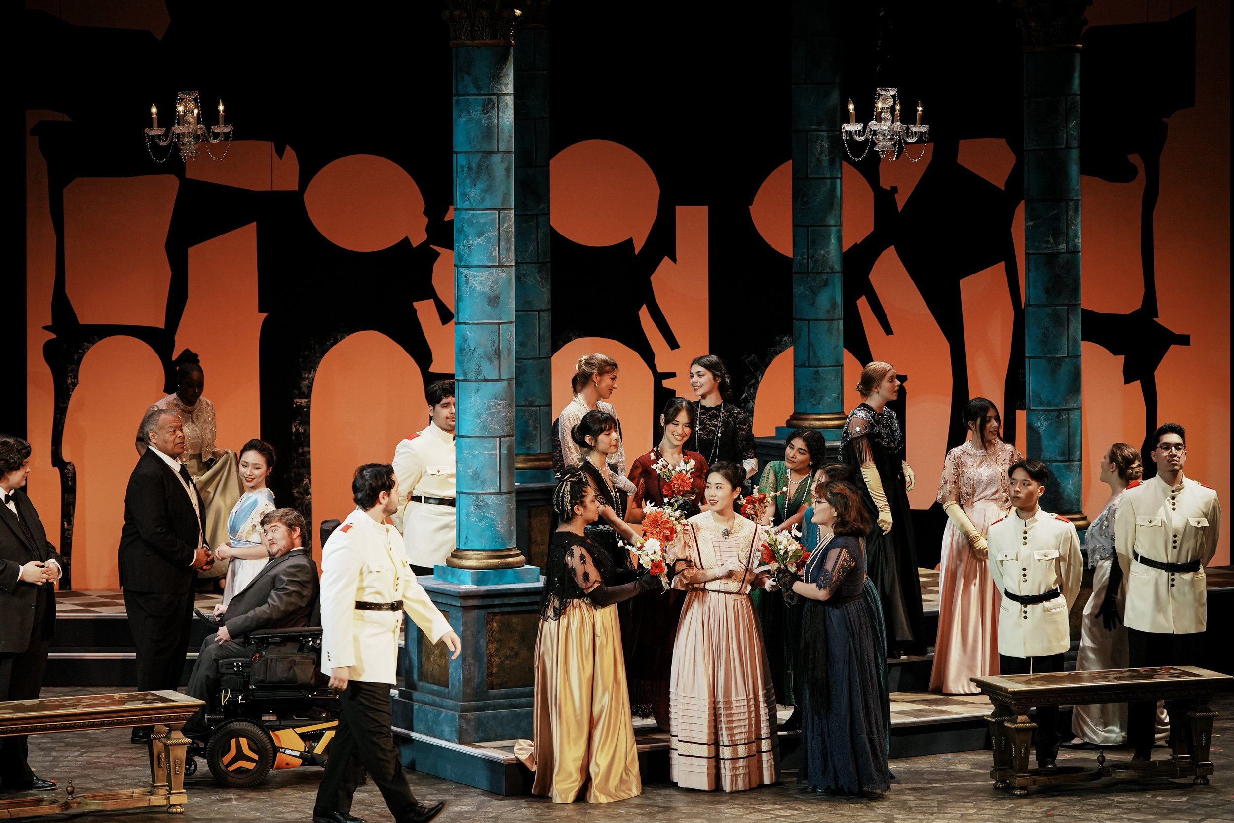 The image shows a theatrical stage with actors in period costumes, including formal dresses and military uniforms, performing on a set designed to resemble a grand interior with tall blue columns and an orange backdrop with black silhouette decoratio