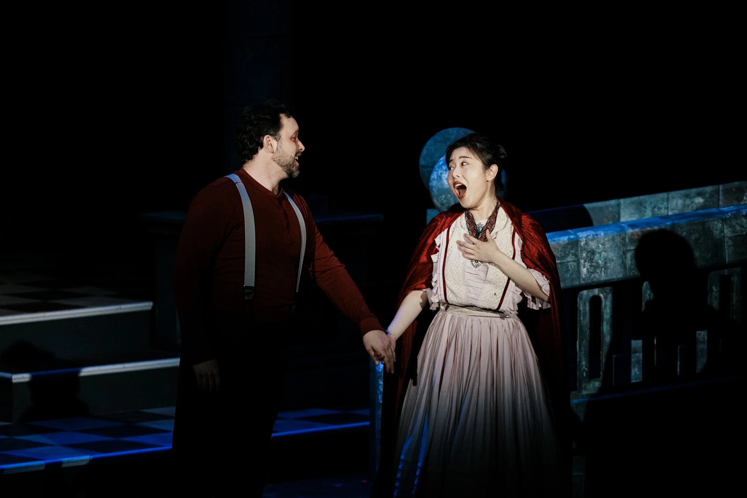 Actors performing on stage, a man and a woman holding hands, the woman with a surprised or emotional expression, stage set with stairs and blue circular decorations.