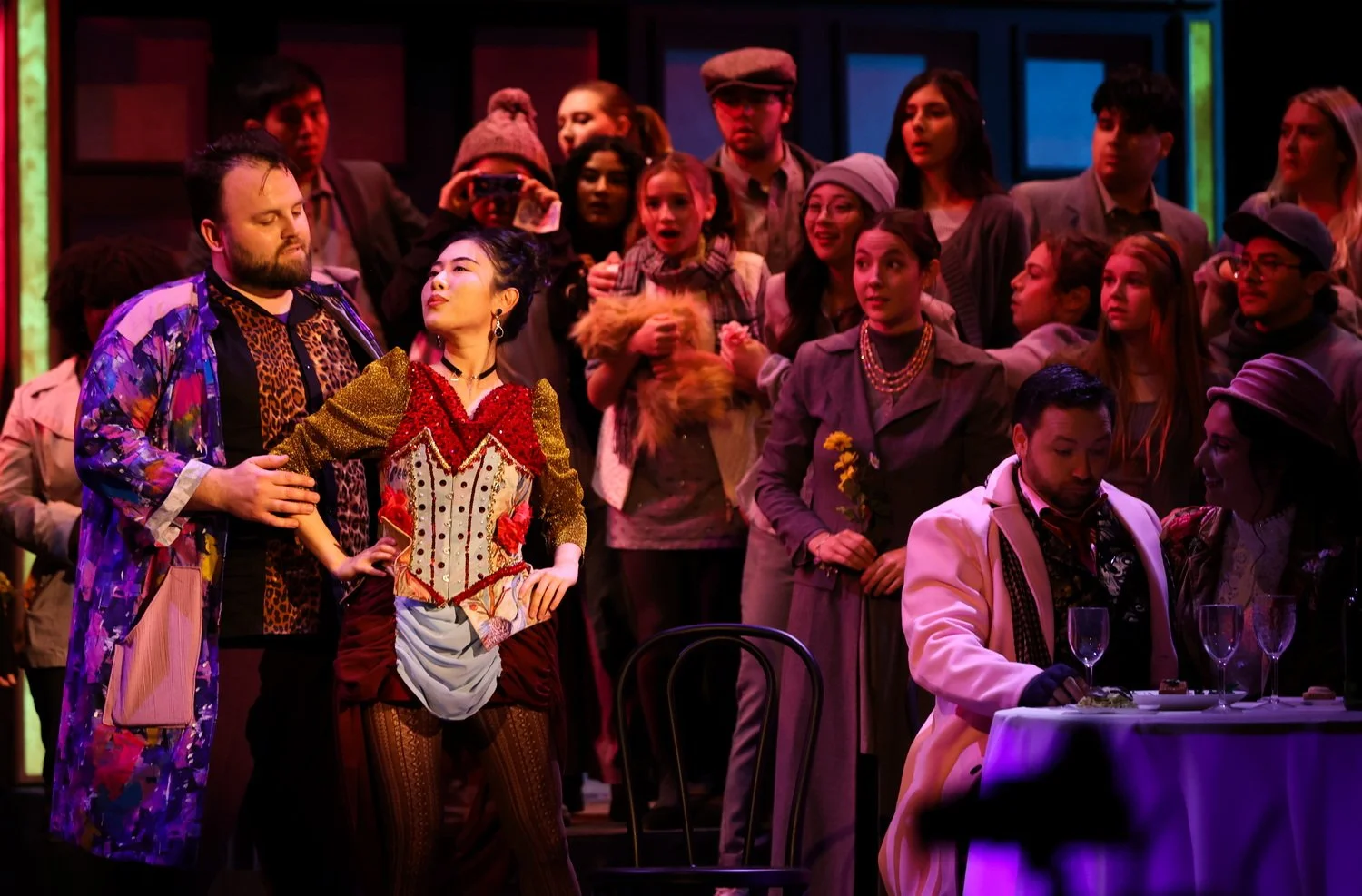Group of actors on stage during a theatrical performance, with some in elaborate costumes and others in everyday clothing, in front of a colorful set.