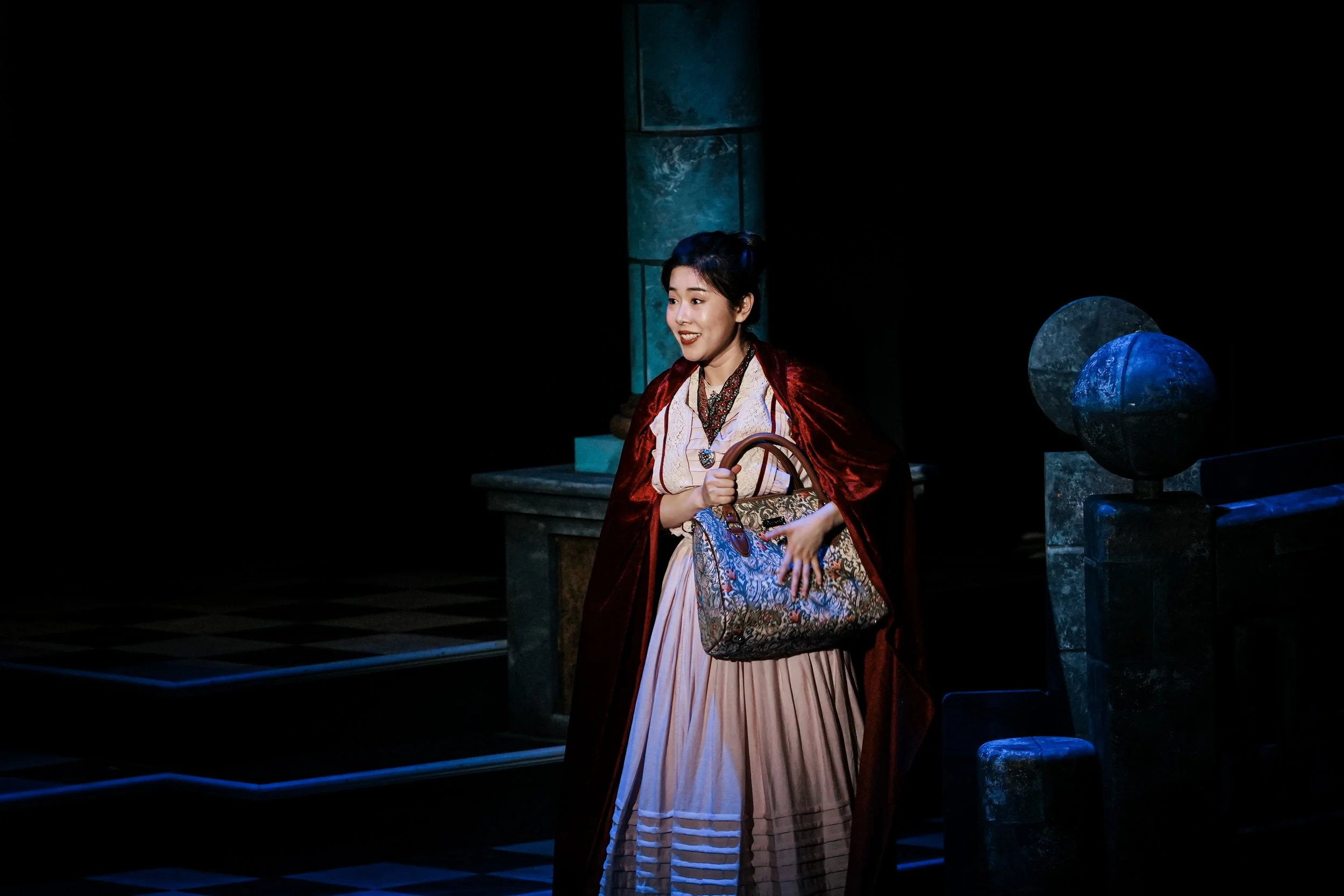 A woman in period costume holding a patterned handbag on a dark stage with stone-like set pieces and blue lighting.
