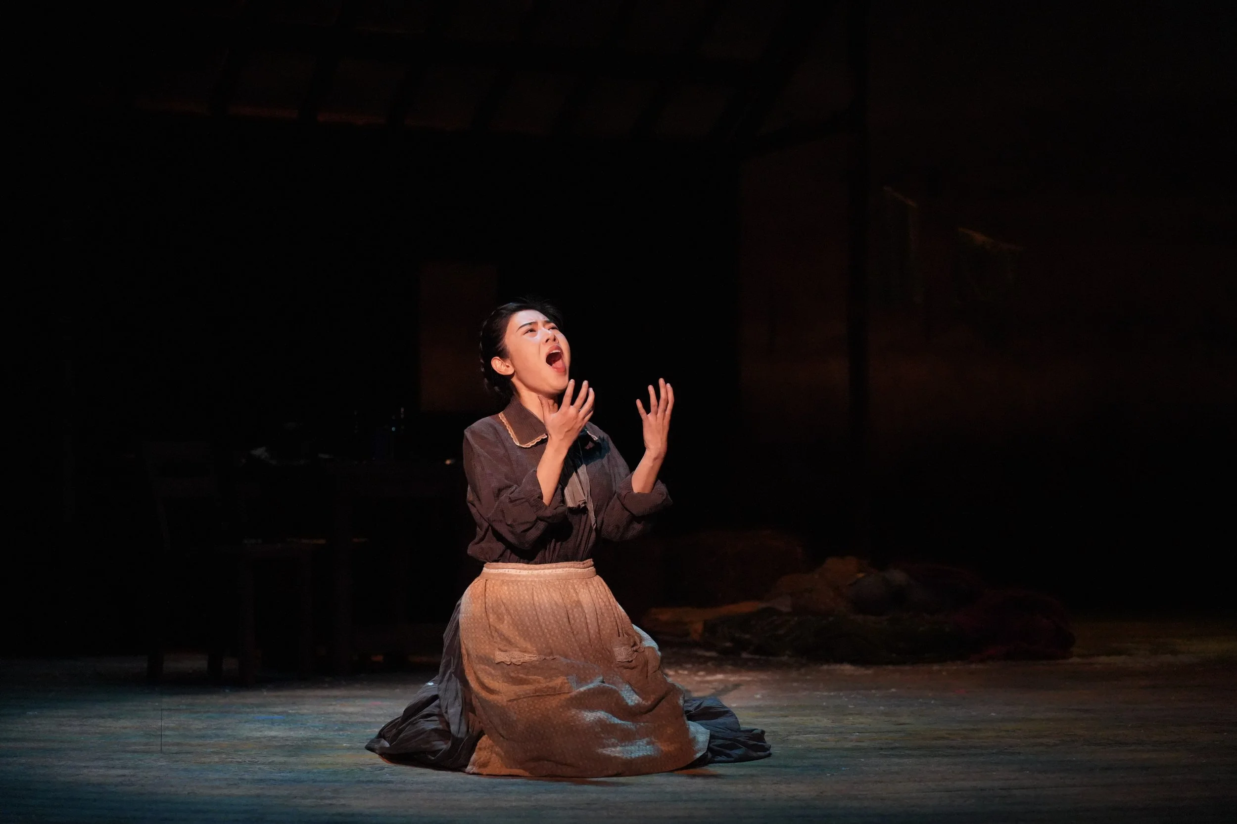 A woman is kneeling on a stage, wearing a dark shirt and a long pleated skirt, passionately singing or acting with her mouth open and hands raised. The background is dark with minimal lighting.