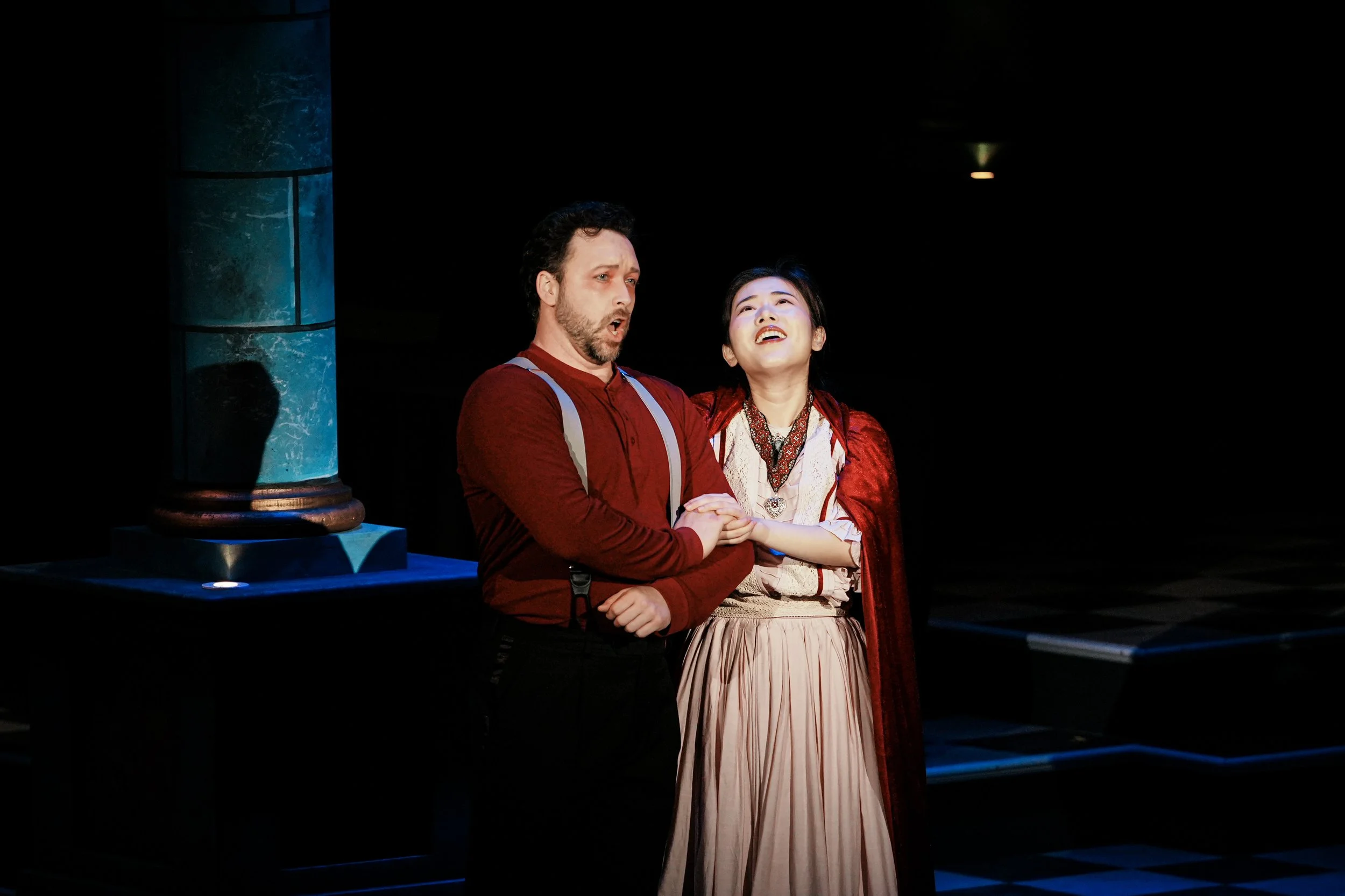 Two actors performing on stage, a man and a woman, with the man appearing emotional and the woman singing or speaking passionately, against a dark background with a blue-colored column.