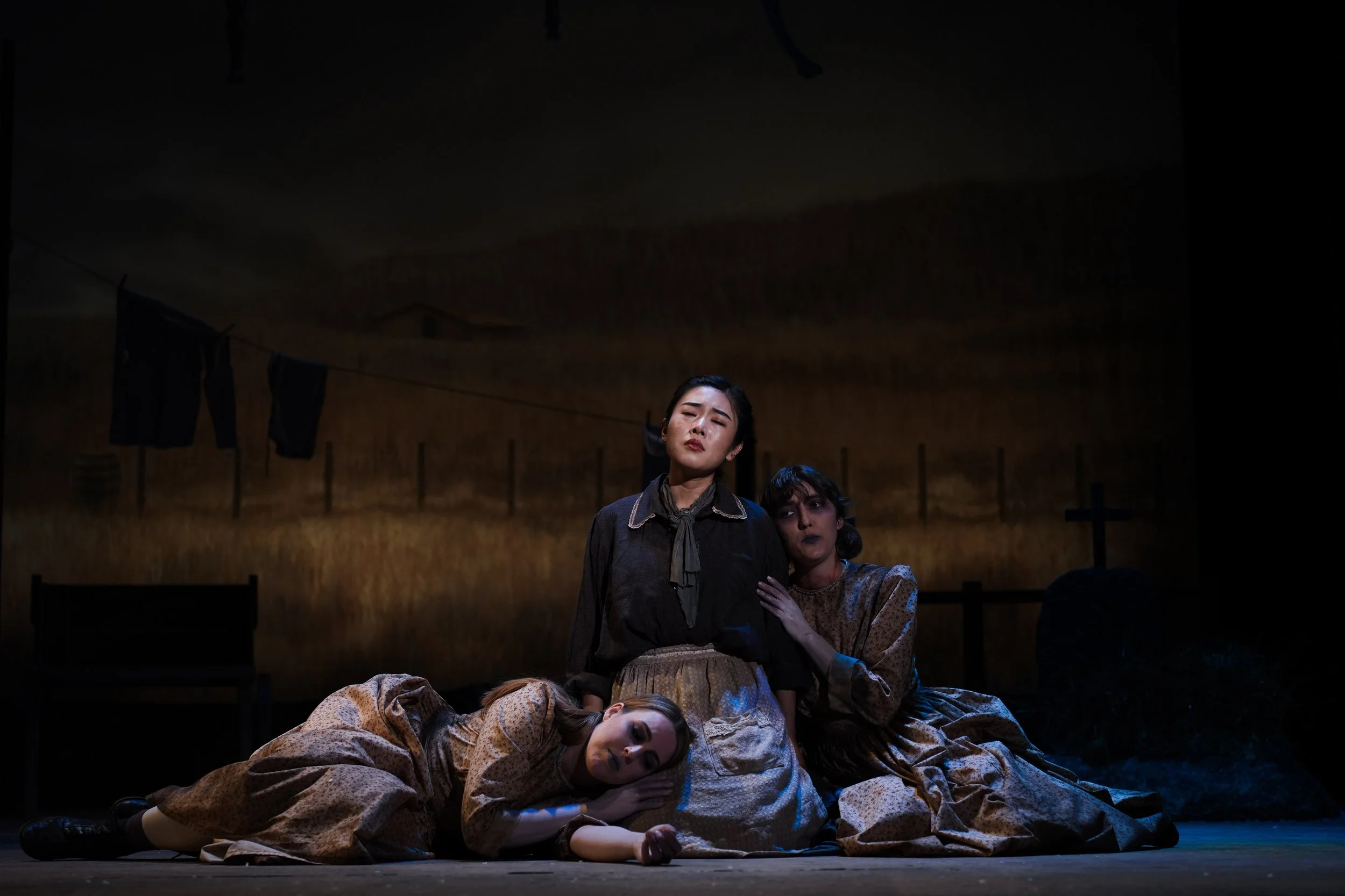 Three women on stage, one sitting and two kneeling, in front of a painted rural landscape backdrop. The woman in the center appears somber, with the other two showing concern or sorrow.