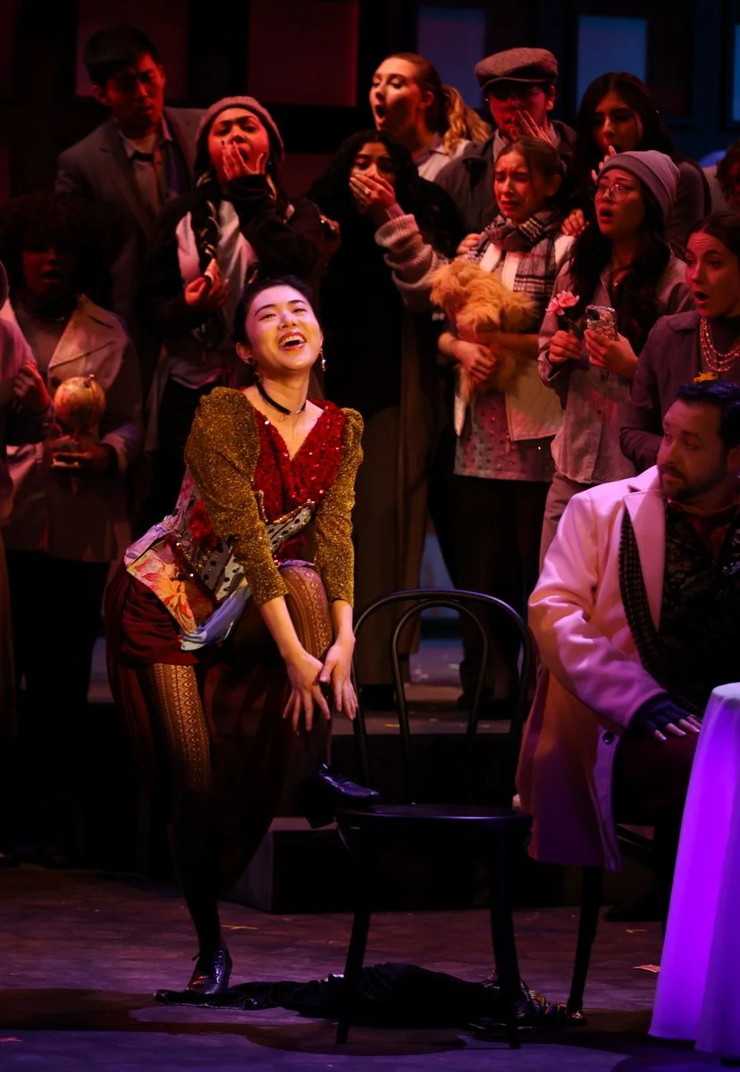 A group of people on stage, some covering their mouths in surprise, with a woman in colorful costume smiling and leaning on a chair, surrounded by others in varied costumes and expressions.