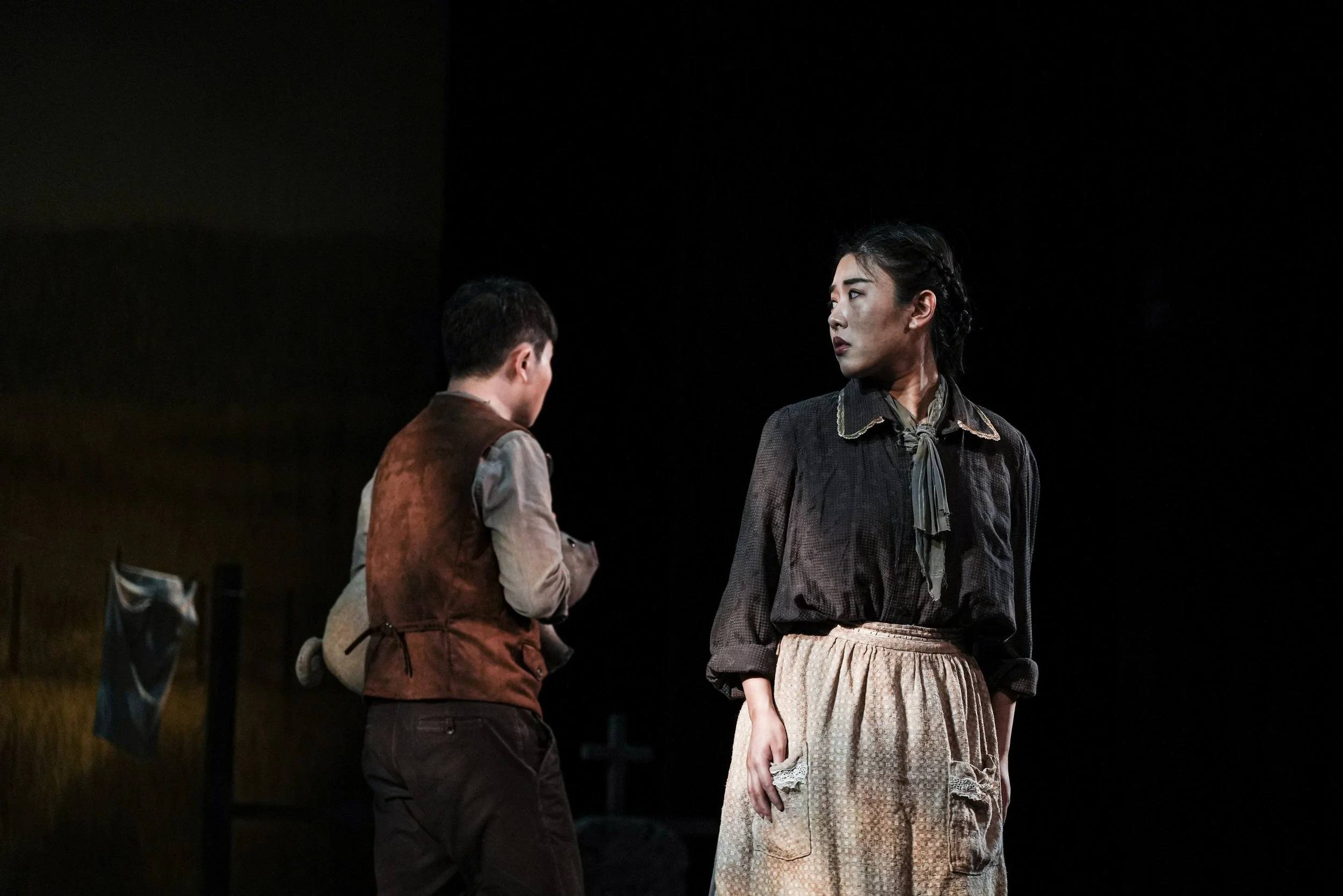 Two actors on stage during a theatrical performance, one with her back to the audience and the other facing her. The woman facing forward is wearing a black blouse with a collar and a beige skirt, and the other actor is in a brown vest and dark pants