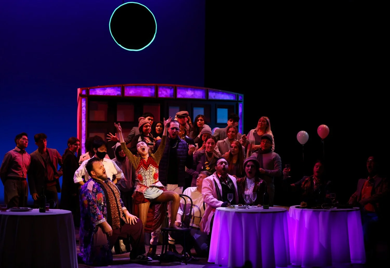 The image depicts a theater stage with a multicultural cast of performers and audience members. The scene appears festive with colorful costumes, balloons, and a vibrant lighting setup, set against a dark background with a glowing circle overhead.