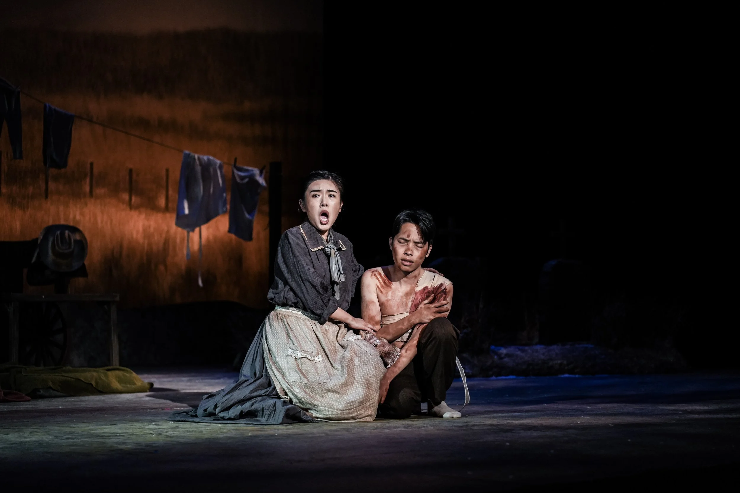 Two actors perform in a theatrical play on stage, with one woman kneeling and appearing distressed, and a man sitting, with bloodstains on his shirt and a pained expression. A clothesline with laundry and a hat are visible in the background.