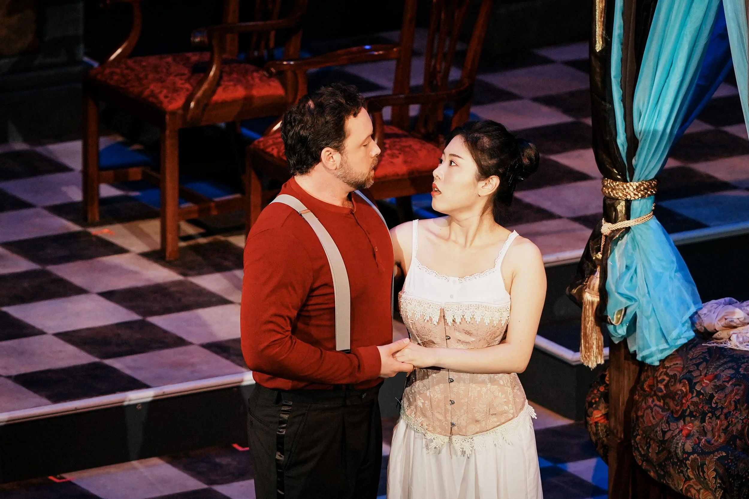 A man and a woman holding hands and looking at each other on a stage with a checkered floor, with chairs and a blue curtain tied back in the background.