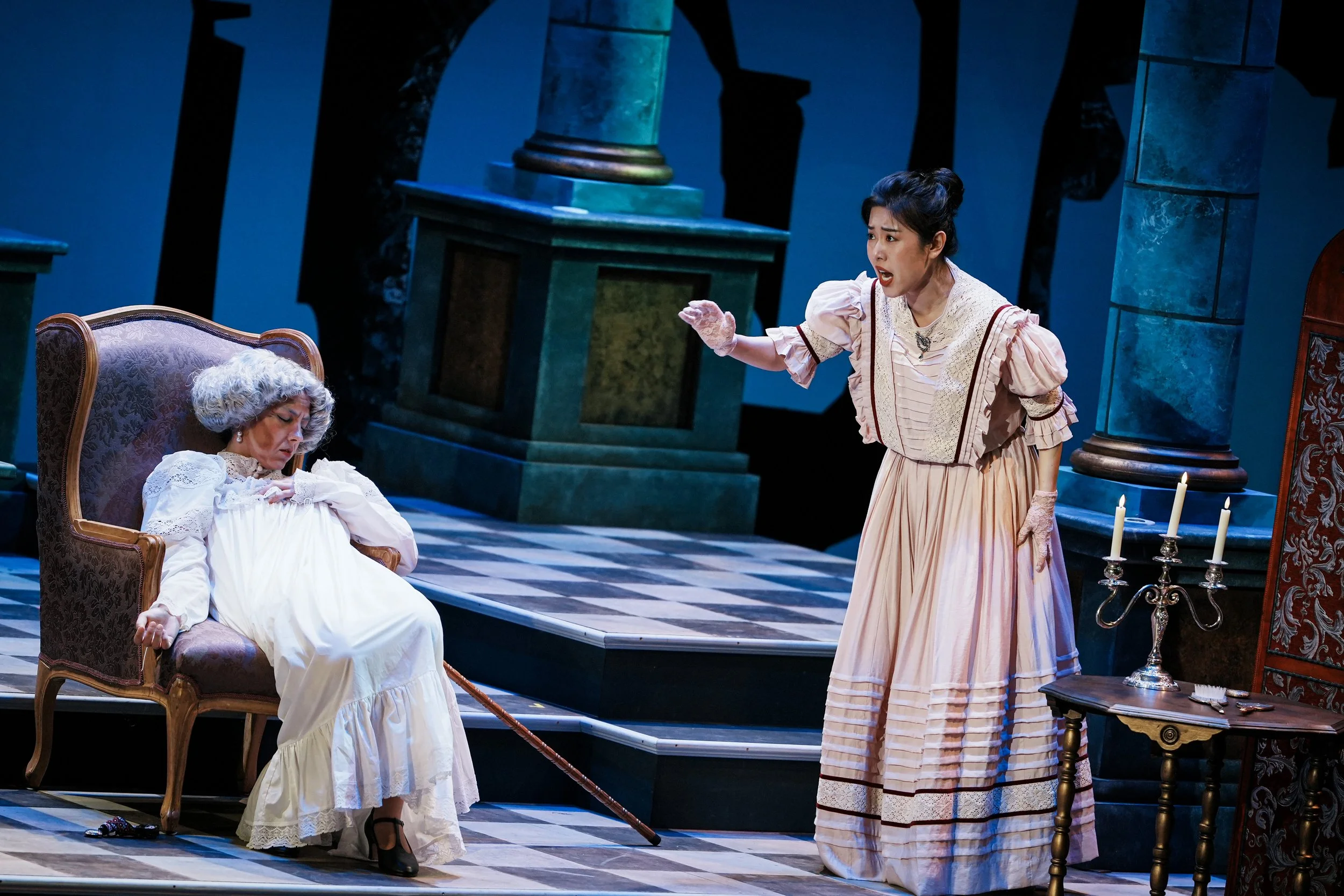 Two women dressed in period costumes are on a stage. One woman with gray hair is sitting in a chair with a cane, appearing upset or drowsy. The other woman, standing, is gesturing and appears to be speaking passionately. The stage has a checkered flo