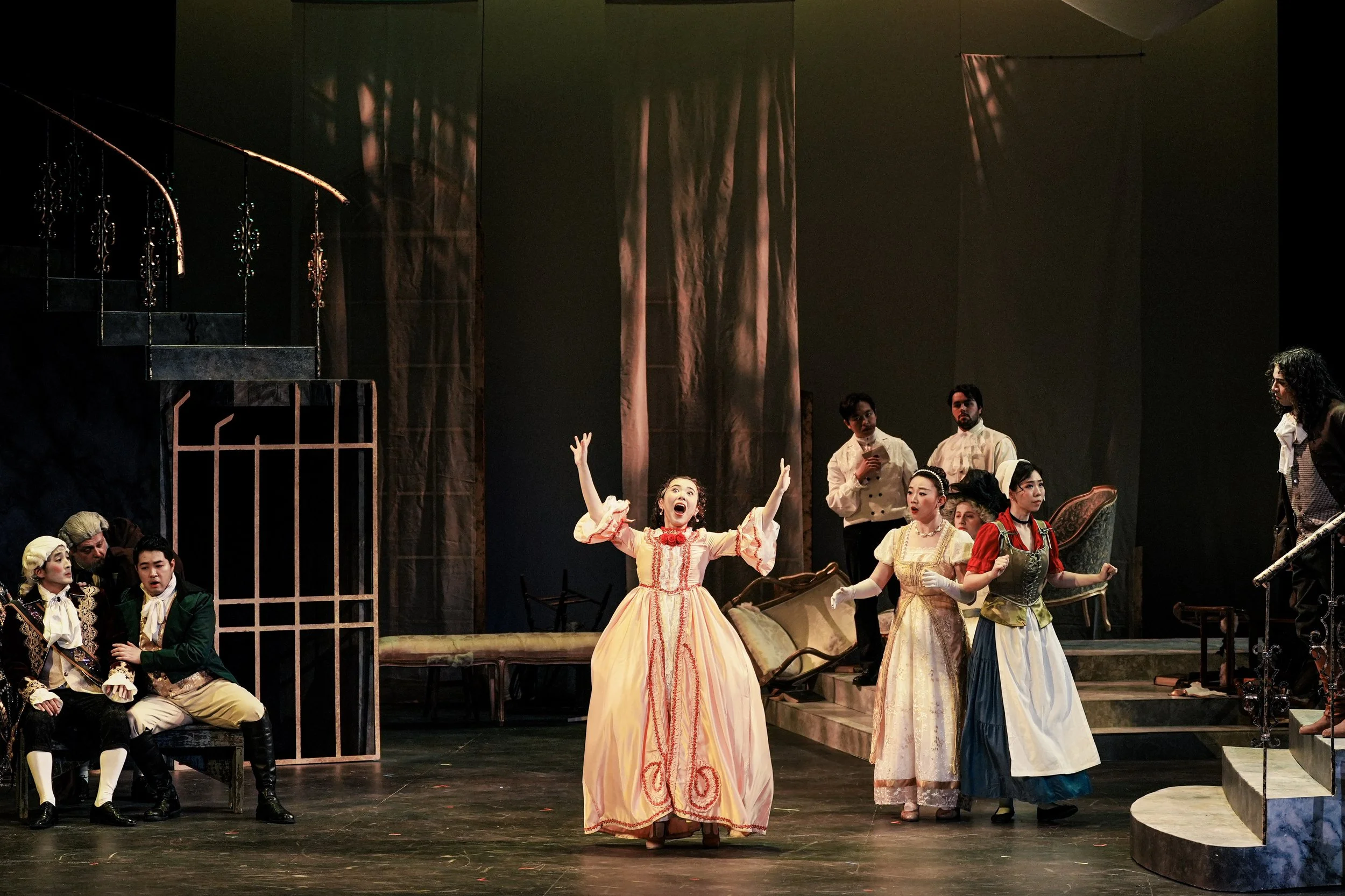 Actors on stage performing a scene from a theatrical play with period costumes, including a woman in a pink gown in the center, surrounded by other characters in vintage attire, on a set with stairs, furniture, and dark background.