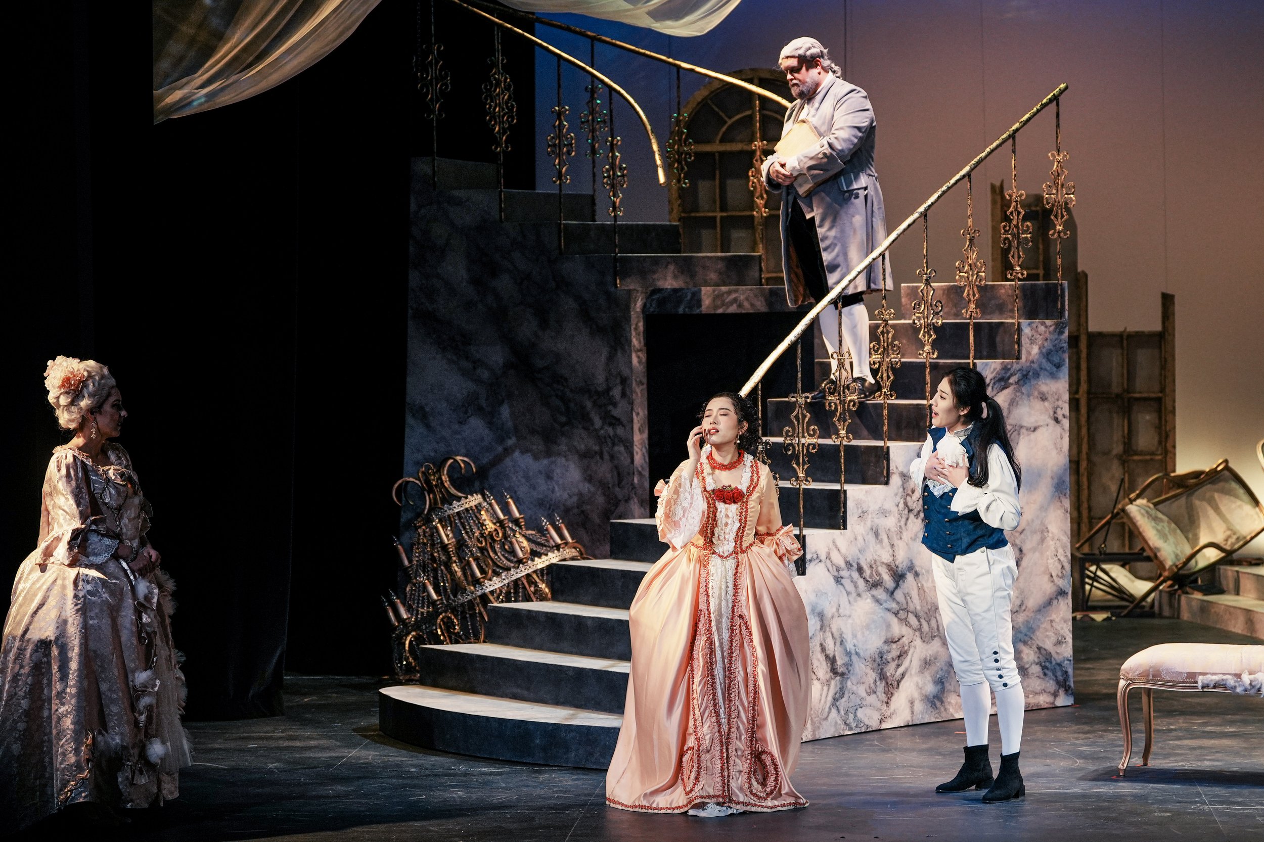 Actors performing a stage play with Victorian costumes and a marble-effect staircase as set decoration.