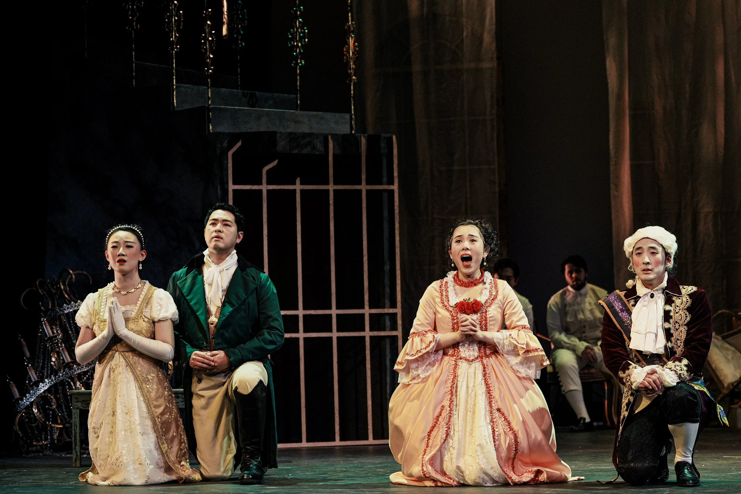 Four actors performing on stage in period costumes, kneeling and singing with solemn expressions, in front of a dark background with minimal set pieces.