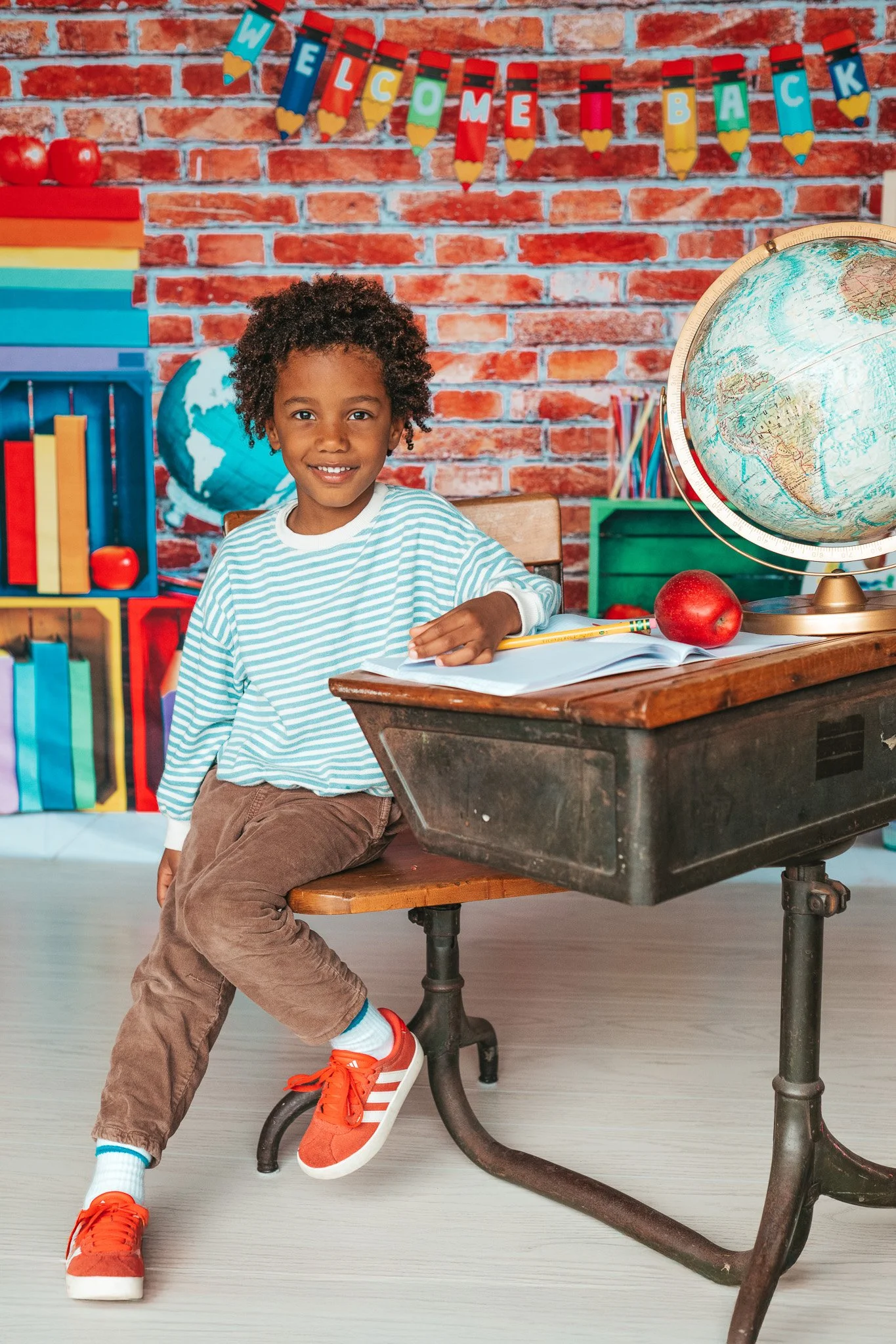 A young boy with curly hair, wearing a striped shirt and brown pants, sitting on a school desk in a colorful classroom with educational decorations and globes, smiling at the camera.