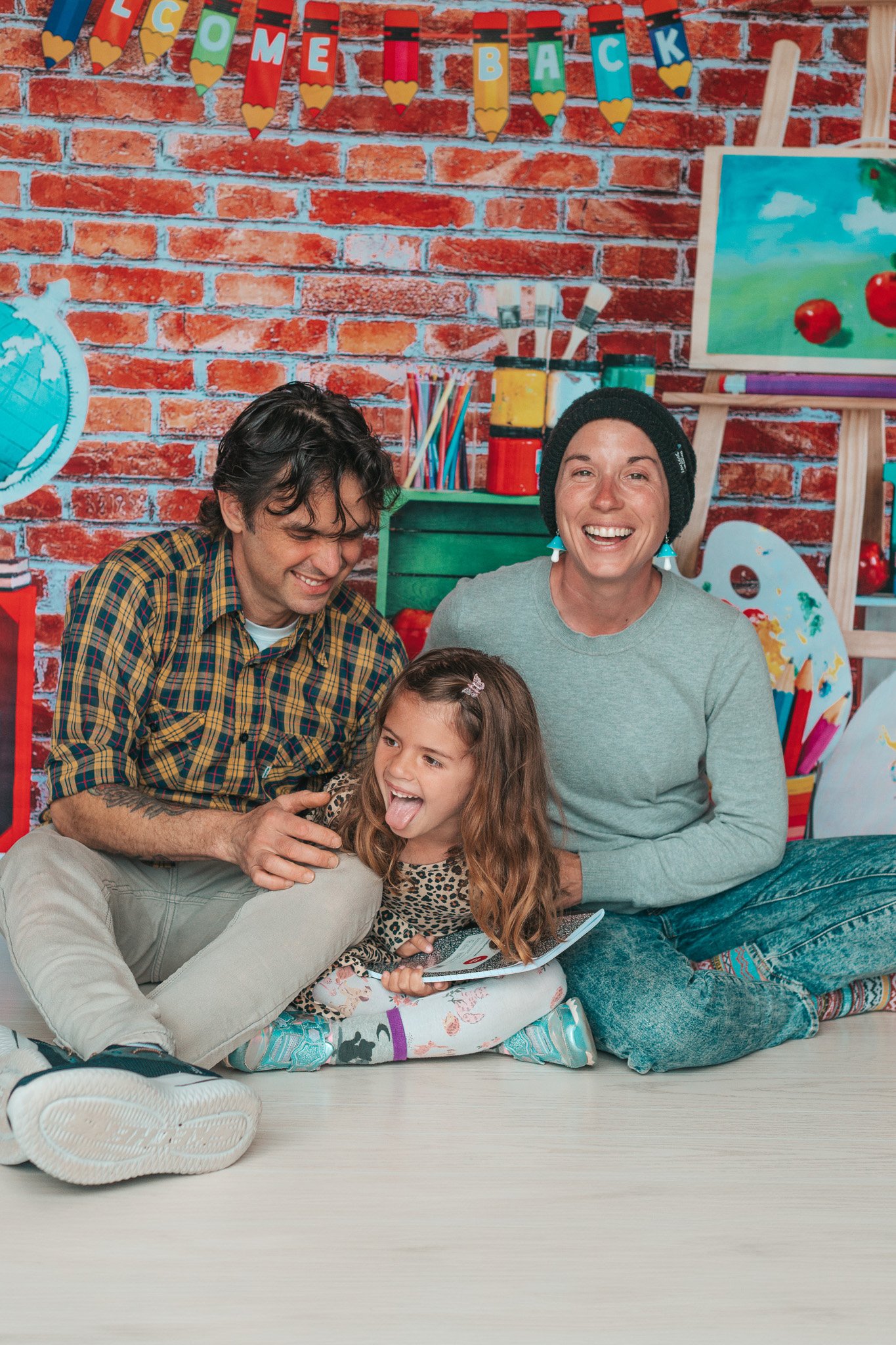 A happy family composed of a man, a woman, and a young girl sitting on the floor together, smiling and laughing, with colorful art supplies and a brick wall in the background.