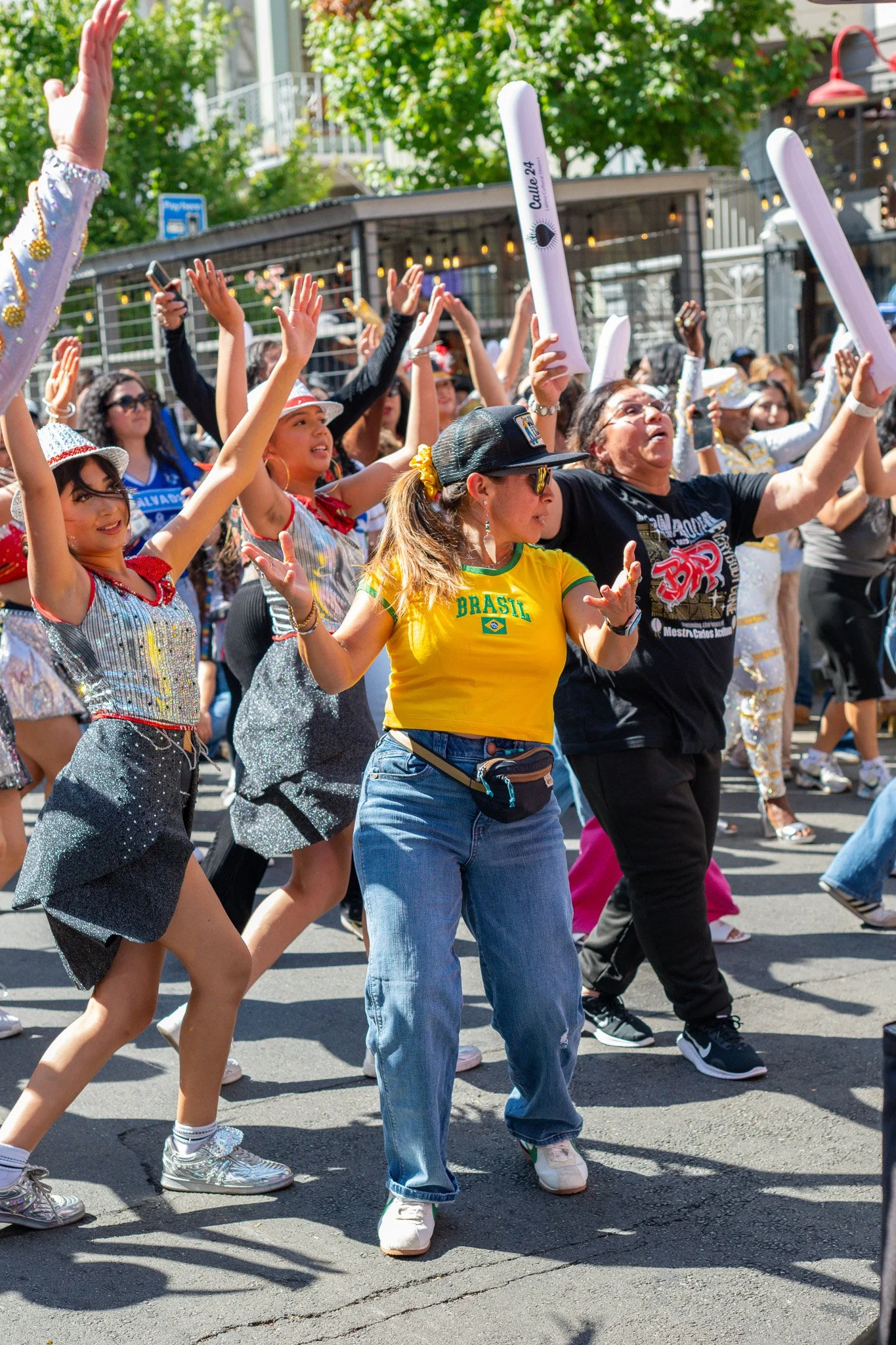 A group of people dancing and celebrating outdoors, some holding inflatable sticks, wearing colorful clothing and accessories.