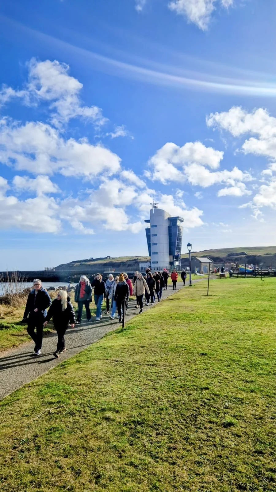 A group of people walking along a walkway near the sea, with a modern white building in the background under a blue sky with clouds.