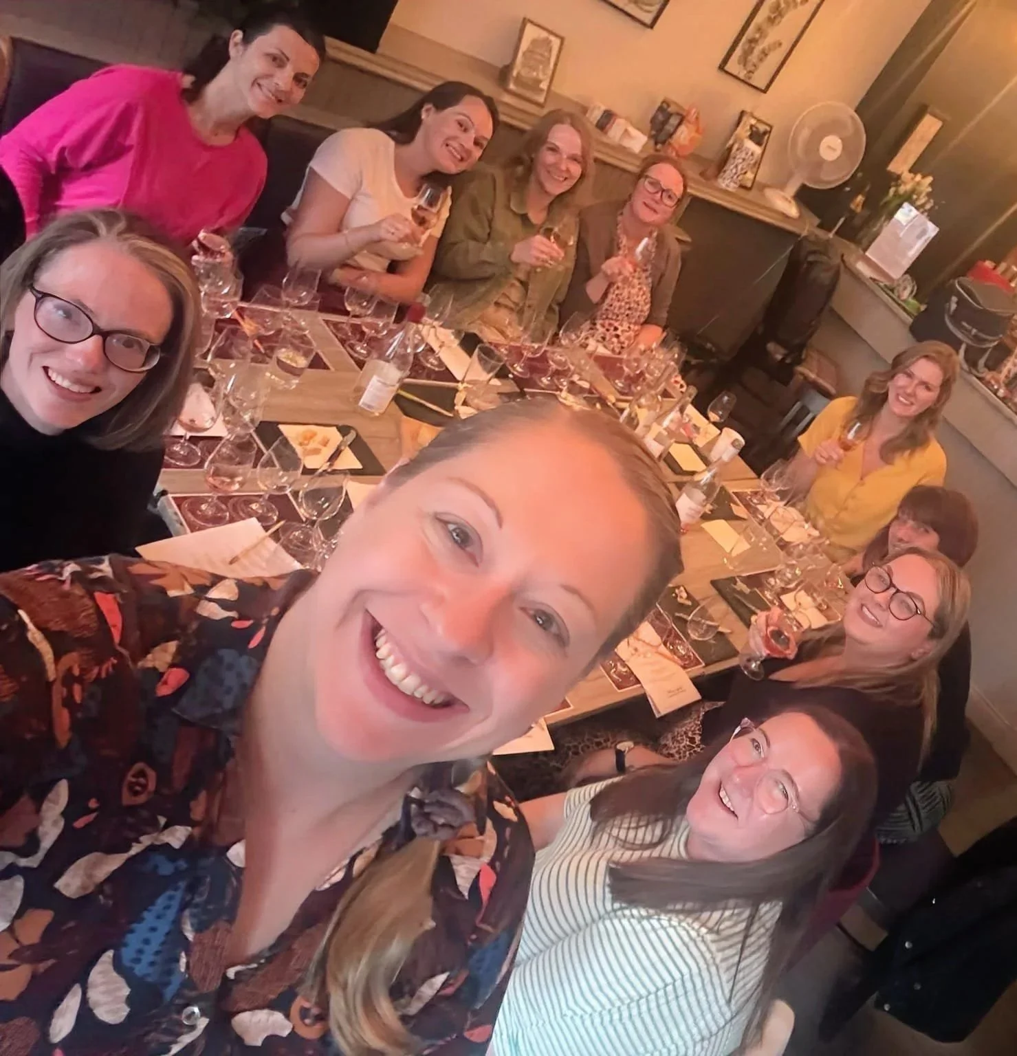 Group of people cheerfully celebrating around a dining table with glasses of wine and food, taking a selfie in a cozy restaurant.