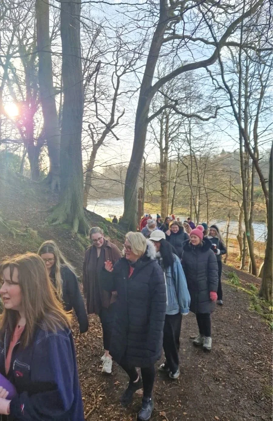 A group of people walking along a wooded trail near a river in a park during late fall or early winter, with some wearing warm clothing.