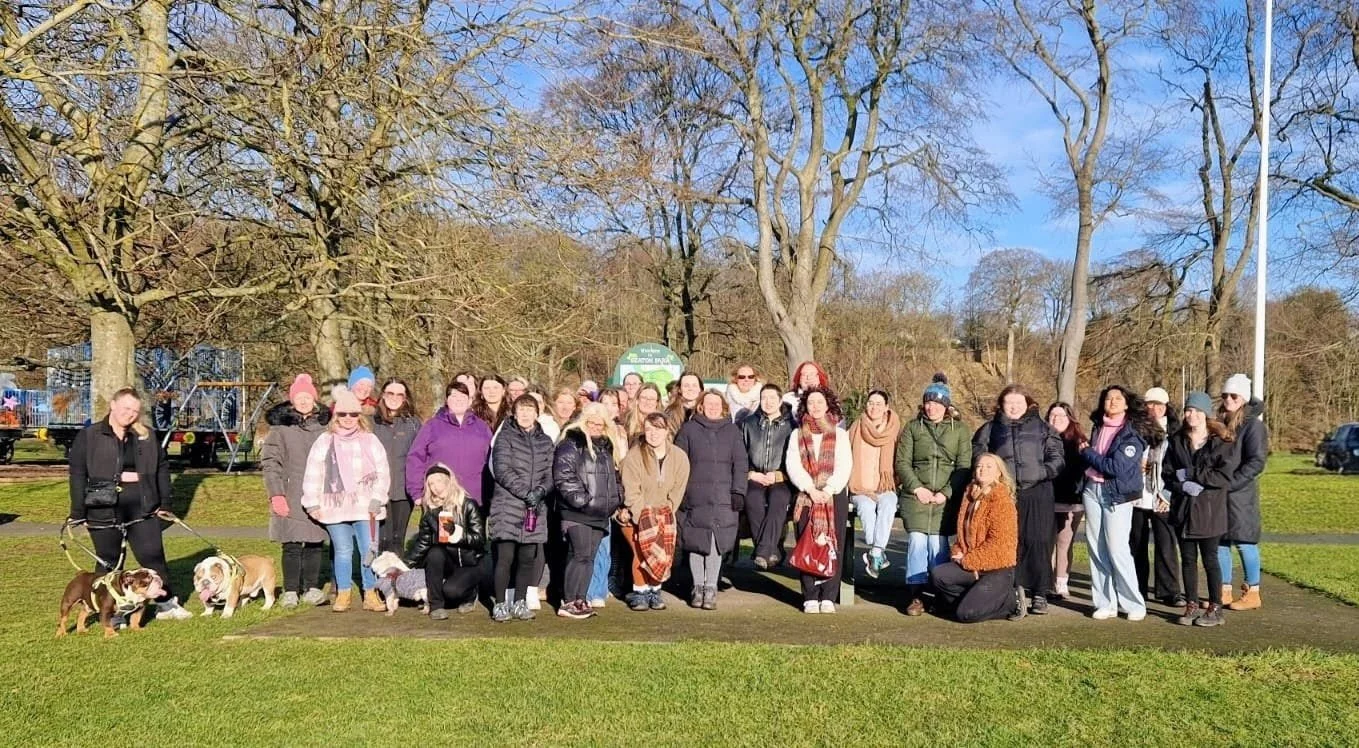 A large group of people standing together outdoors in a park, with some people holding dogs on leashes, during a sunny day with trees in the background.