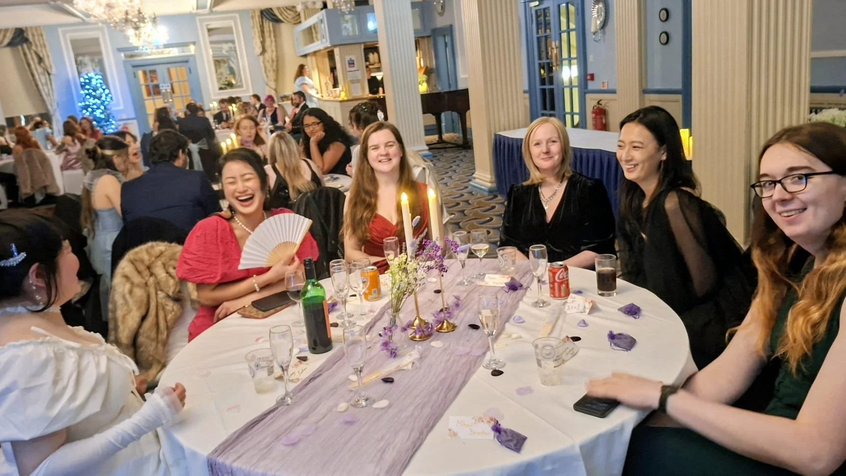 A group of women sitting around a decorated table at a celebration, smiling and enjoying the event. The table has purple and white decorations, flower arrangements, candles, and drinks. In the background, other guests and a decorated Christmas tree are visible.