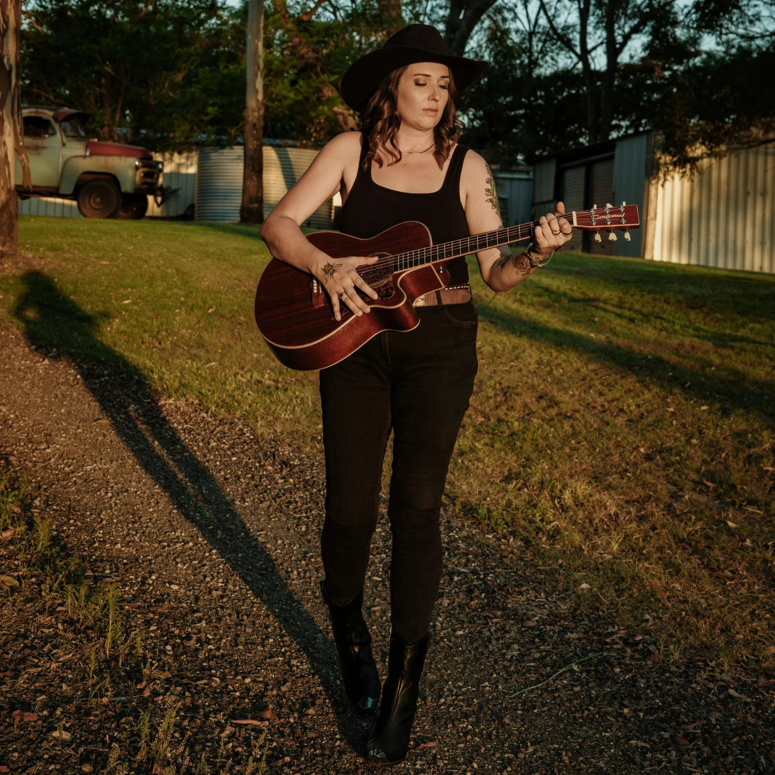 A woman with dark brown hair, wearing a black wide-brim hat, black tank top, black pants, and black boots, playing an acoustic guitar outdoors during sunset.