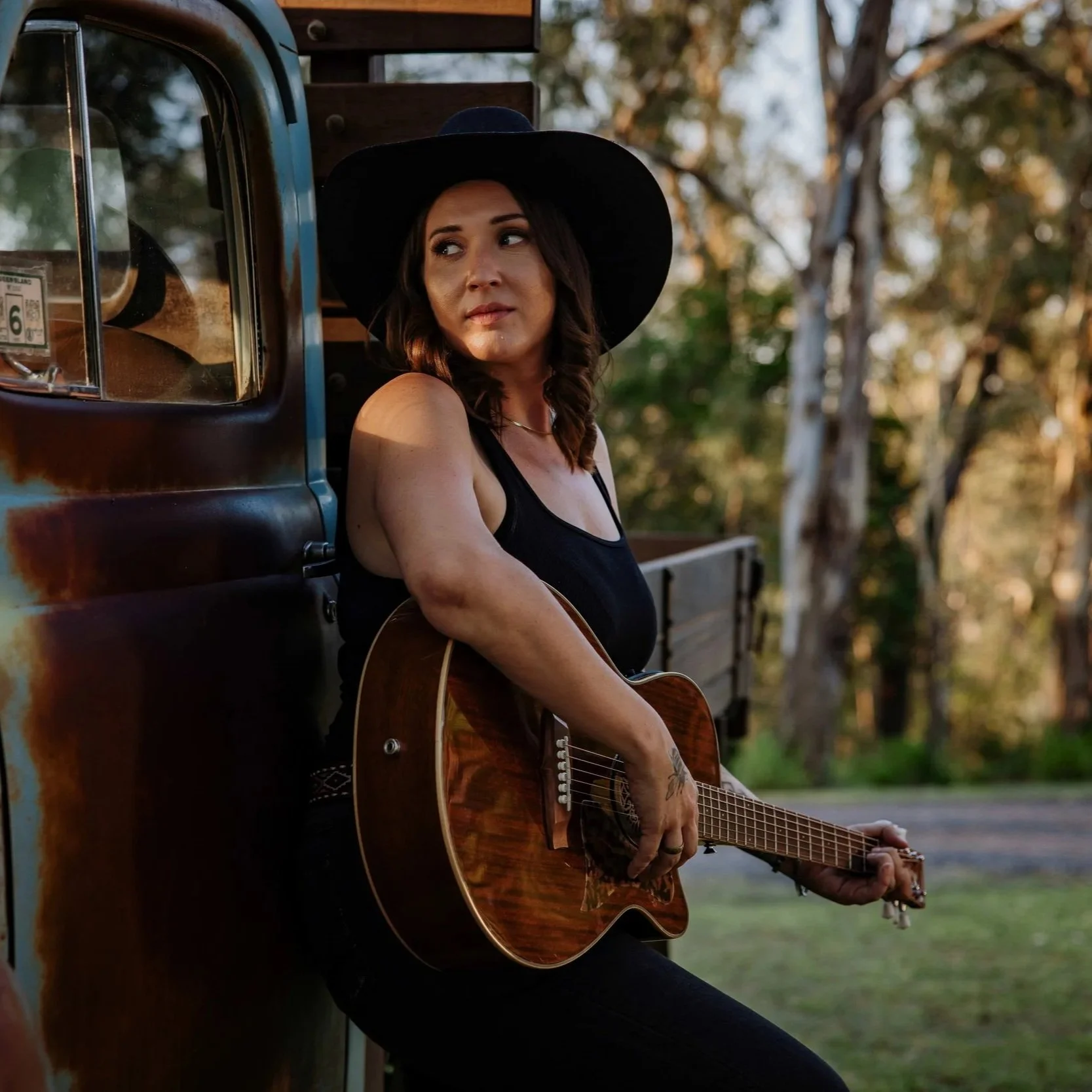 A woman wearing a black sleeveless top and large black hat is sitting beside a weathered green and brown truck, holding an acoustic guitar, in a natural outdoor setting with trees in the background.