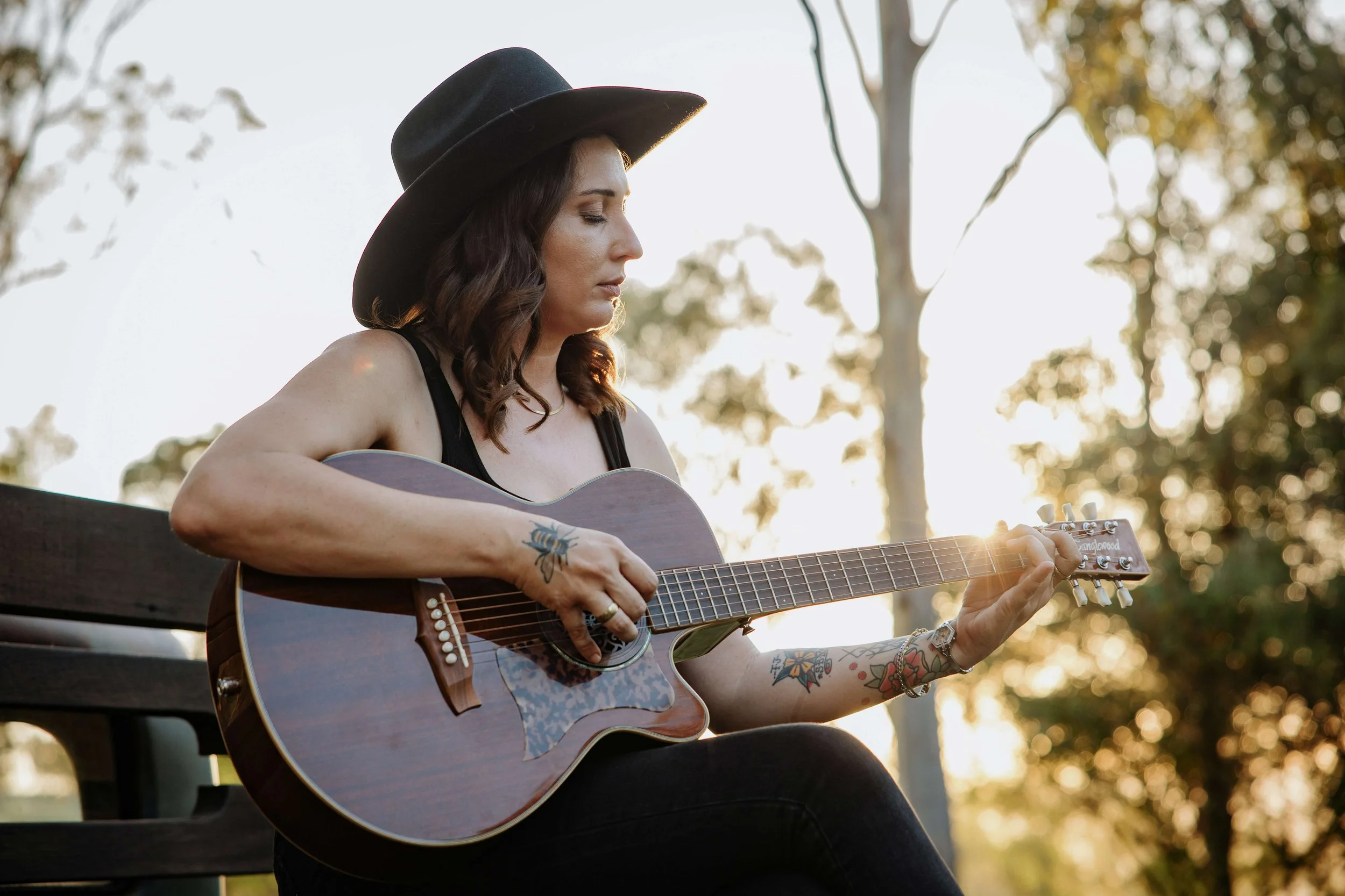 Woman with dark hair wearing a black hat and black tank top playing an acoustic guitar outdoors during sunset.