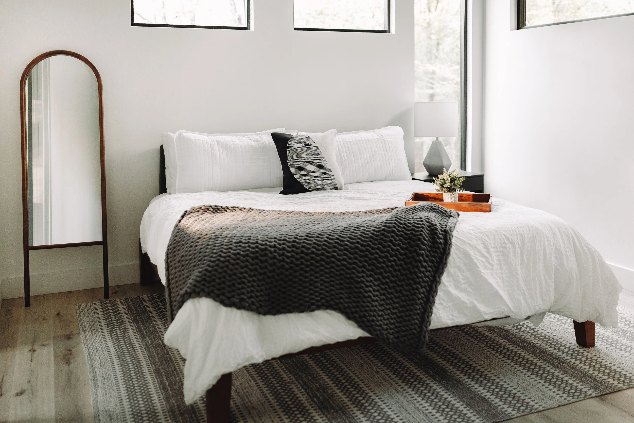 A neatly made bed with white bedding, black and patterned pillows, a chunky gray blanket, and a wooden tray with a small plant on it, beside a bedside table with a lamp, in a modern bedroom with large windows.