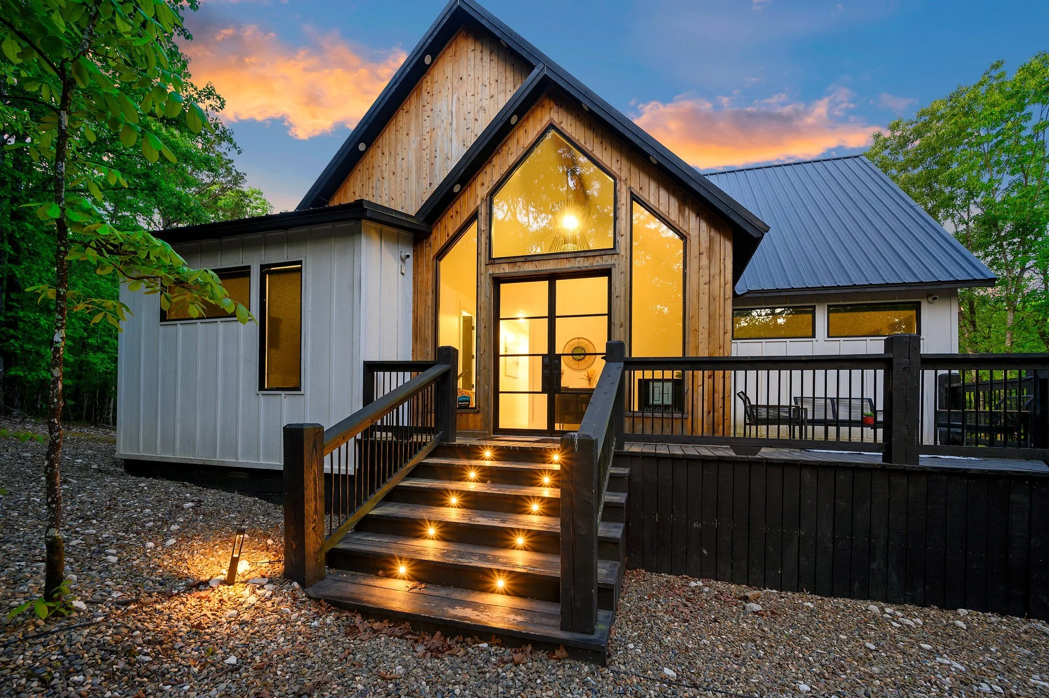 Hygge Hill, a modern cabin with large glass windows and a wooden facade, surrounded by trees during sunset, with illuminated steps leading up to the entrance.