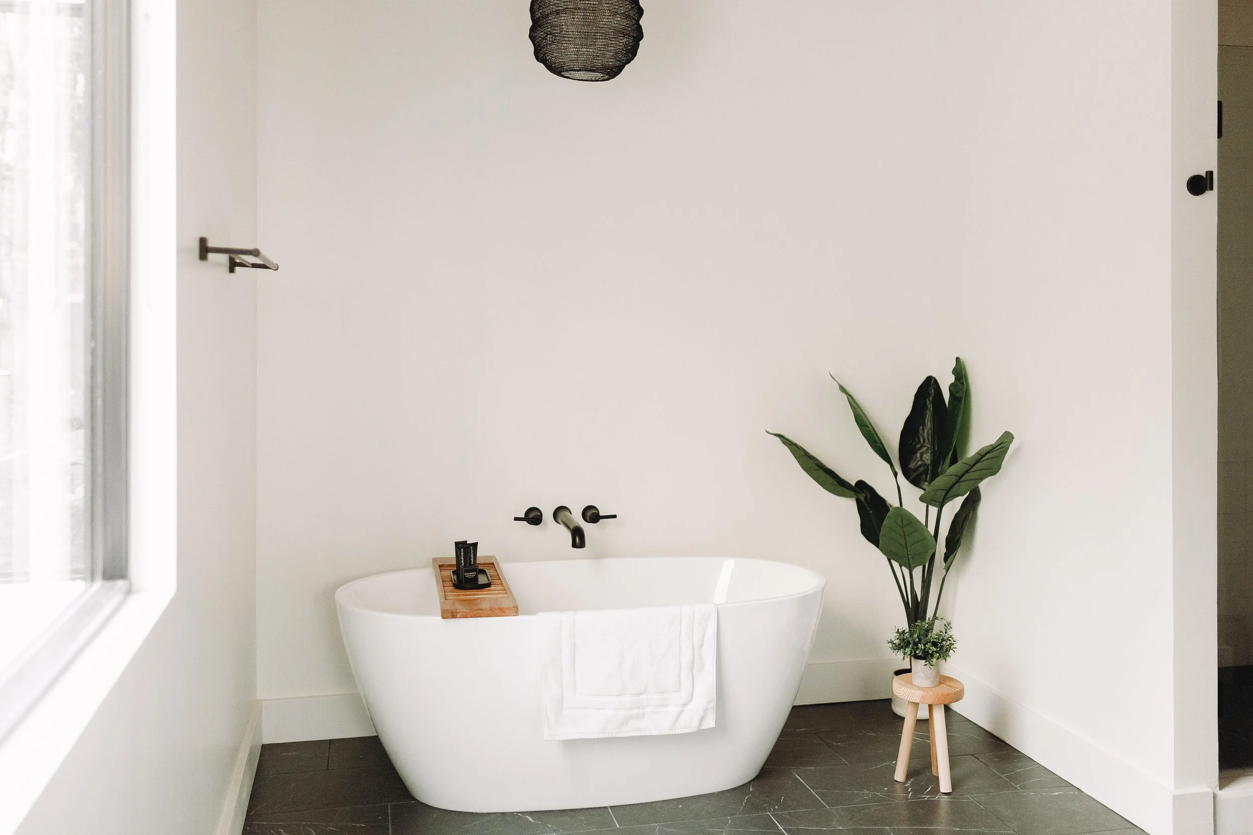 Modern white bathtub with a towel, a wooden tray with toiletries, a black-finish faucet, and a tall green plant in a pot, in a minimalistic bathroom with white walls and black tile flooring.