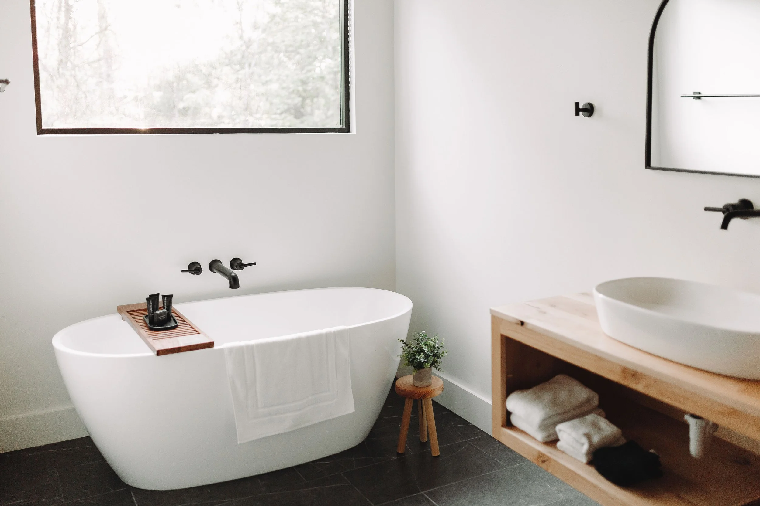 Modern bathroom with a white bathtub, small wooden stool with a potted plant, wooden vanity with towels, and a window letting in natural light.