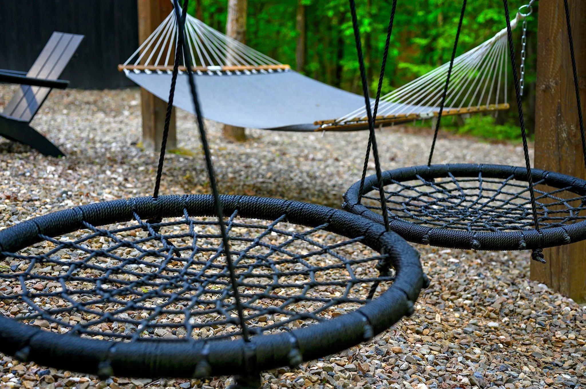 Two black circular net swings hanging from ropes on a gravel ground, with a hammock in the background among trees in a park or backyard.