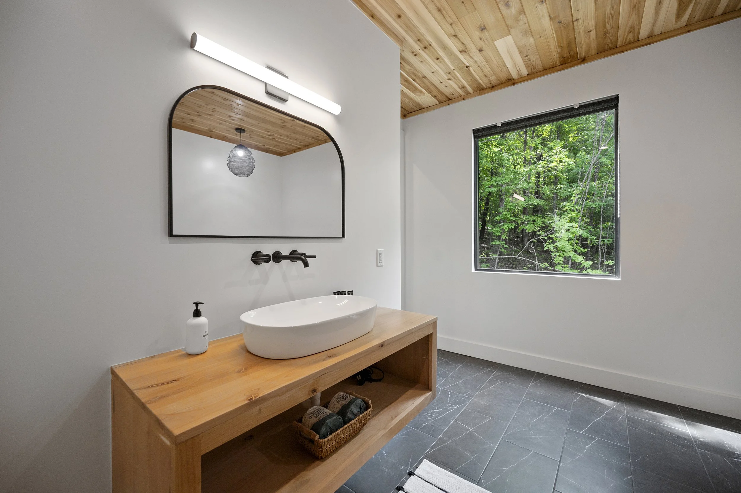 Modern bathroom with a wooden vanity, backsplash, and a vessel sink, large window showing a green forest, and a mirror above the sink.