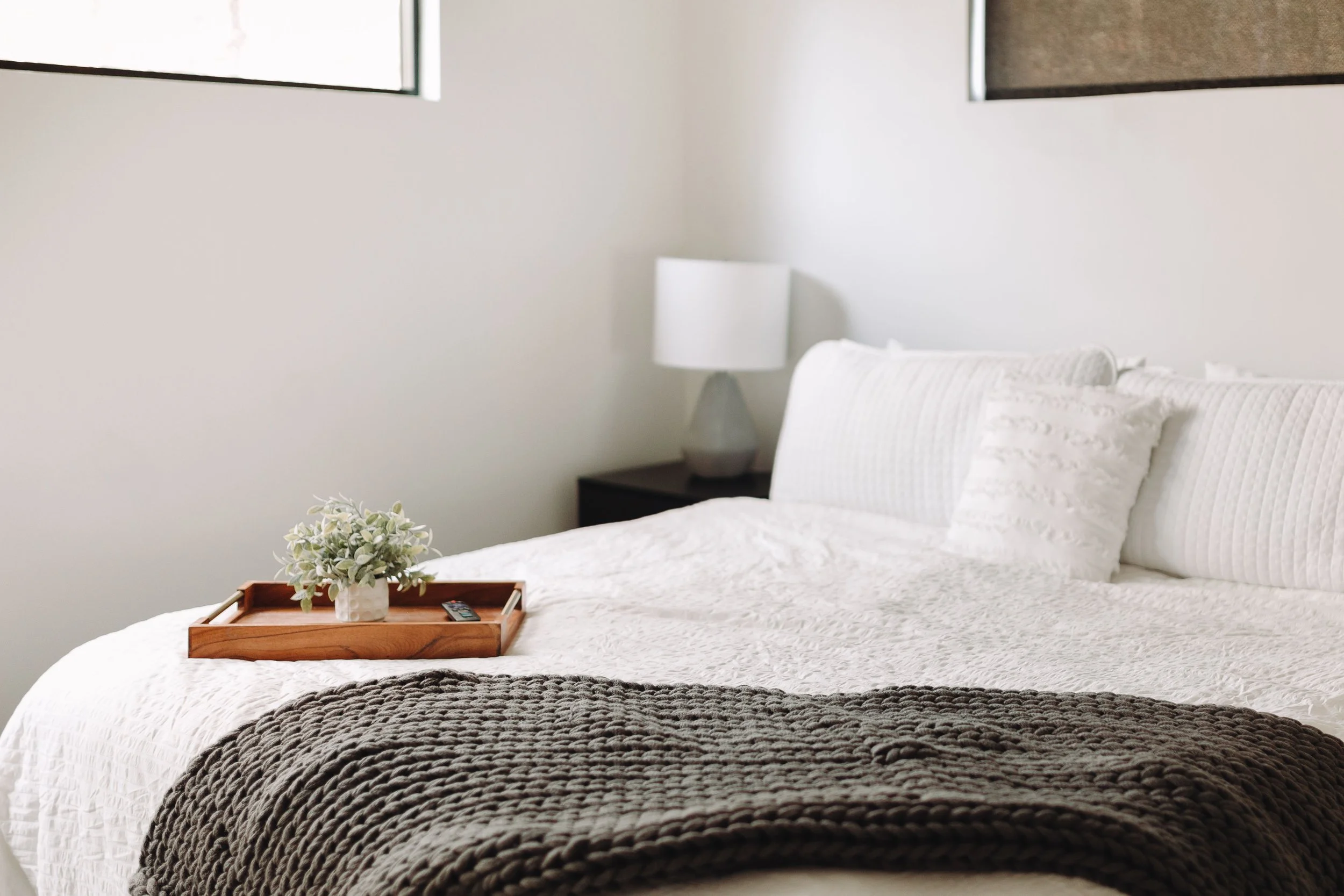 A neatly made bed with white bedding and pillows, a brown knitted throw blanket at the foot, a tray with a potted plant and remote on the bed, a bedside table with a white lamp, and two small windows above the bed, in a minimalistic bedroom.