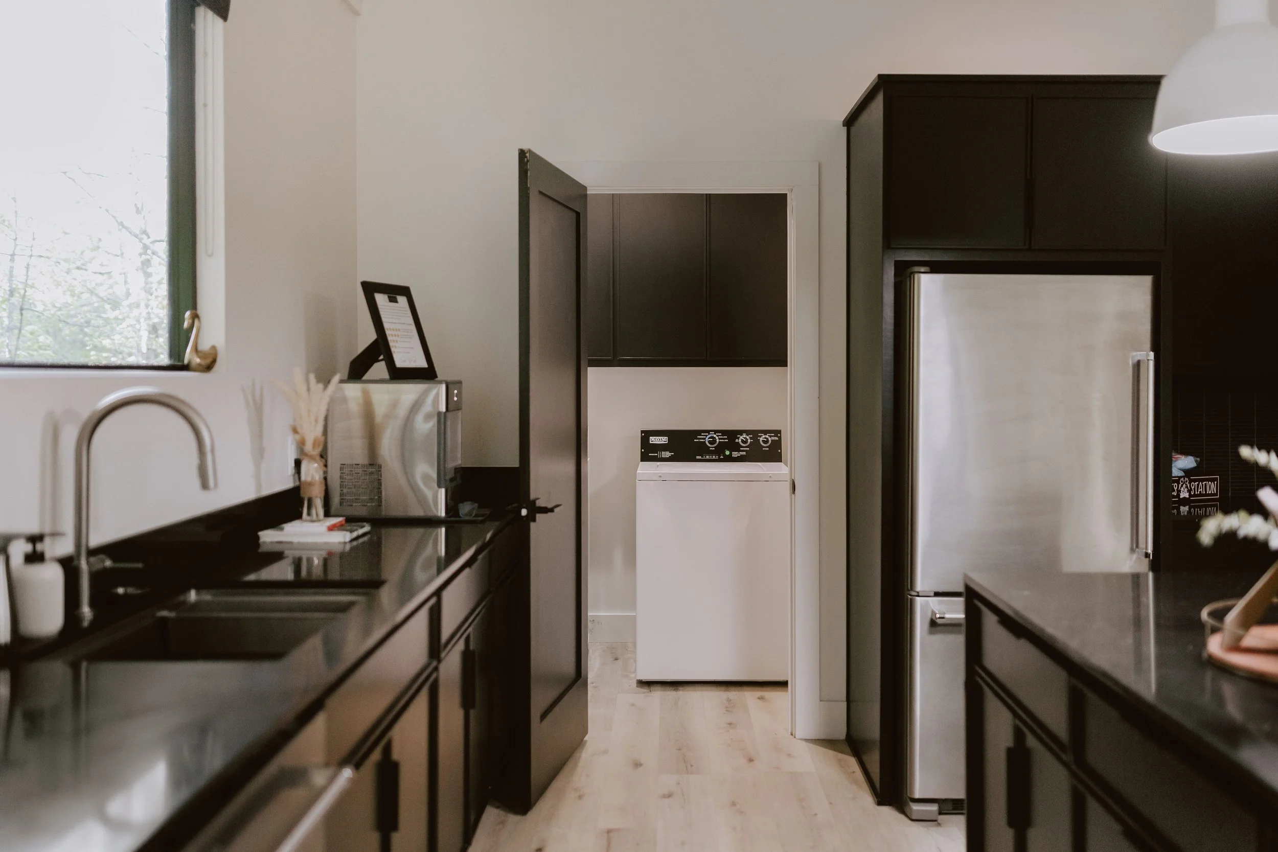Kitchen with black cabinets, stainless steel refrigerator, and a small laundry area with a white washer, black cabinets, and a window with a view of trees outside.