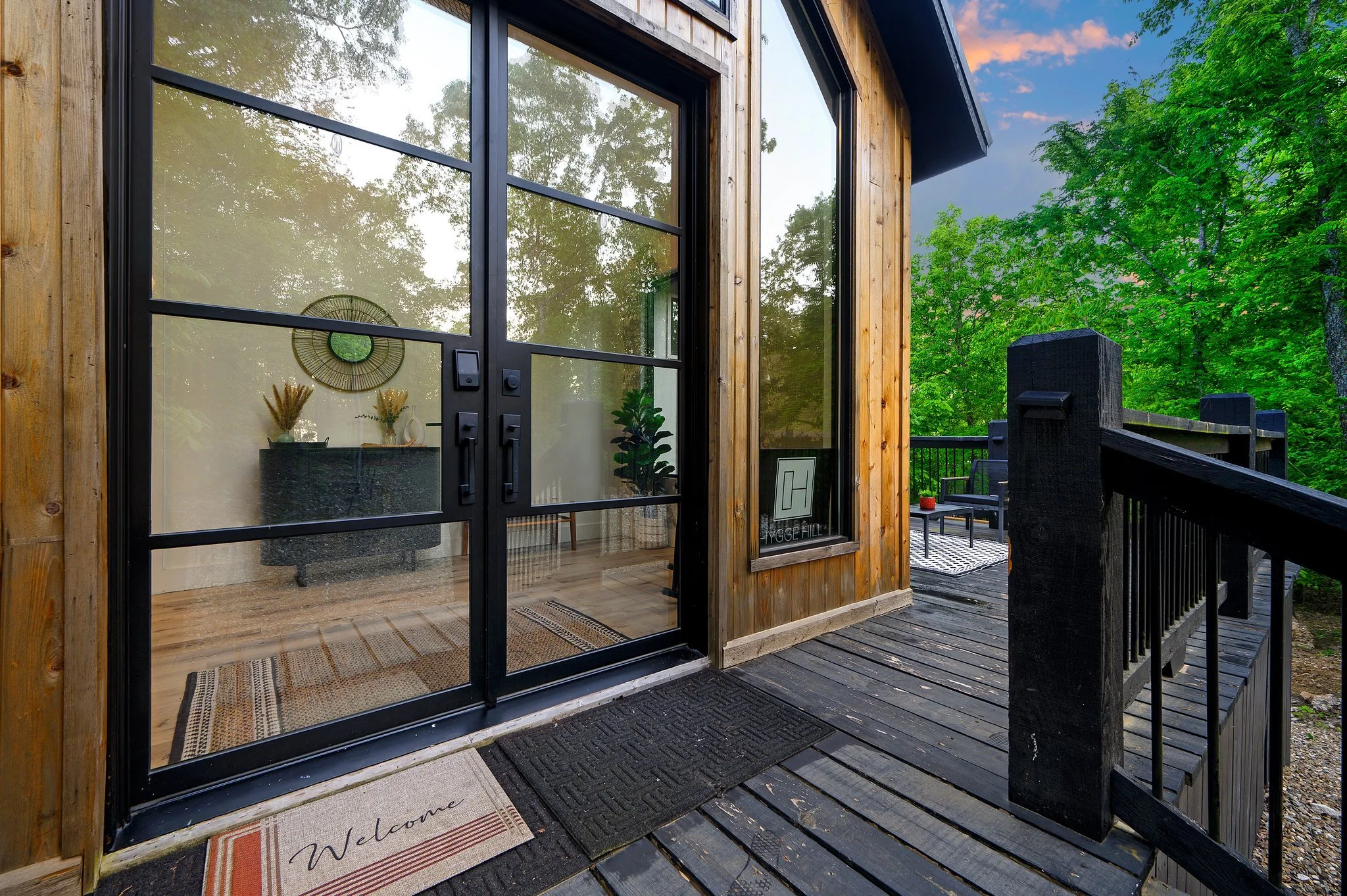 Front porch of Hygge Hill, a modern cabin, with a black door and large glass windows, surrounded by greenery, with a small outdoor seating area visible to the right.