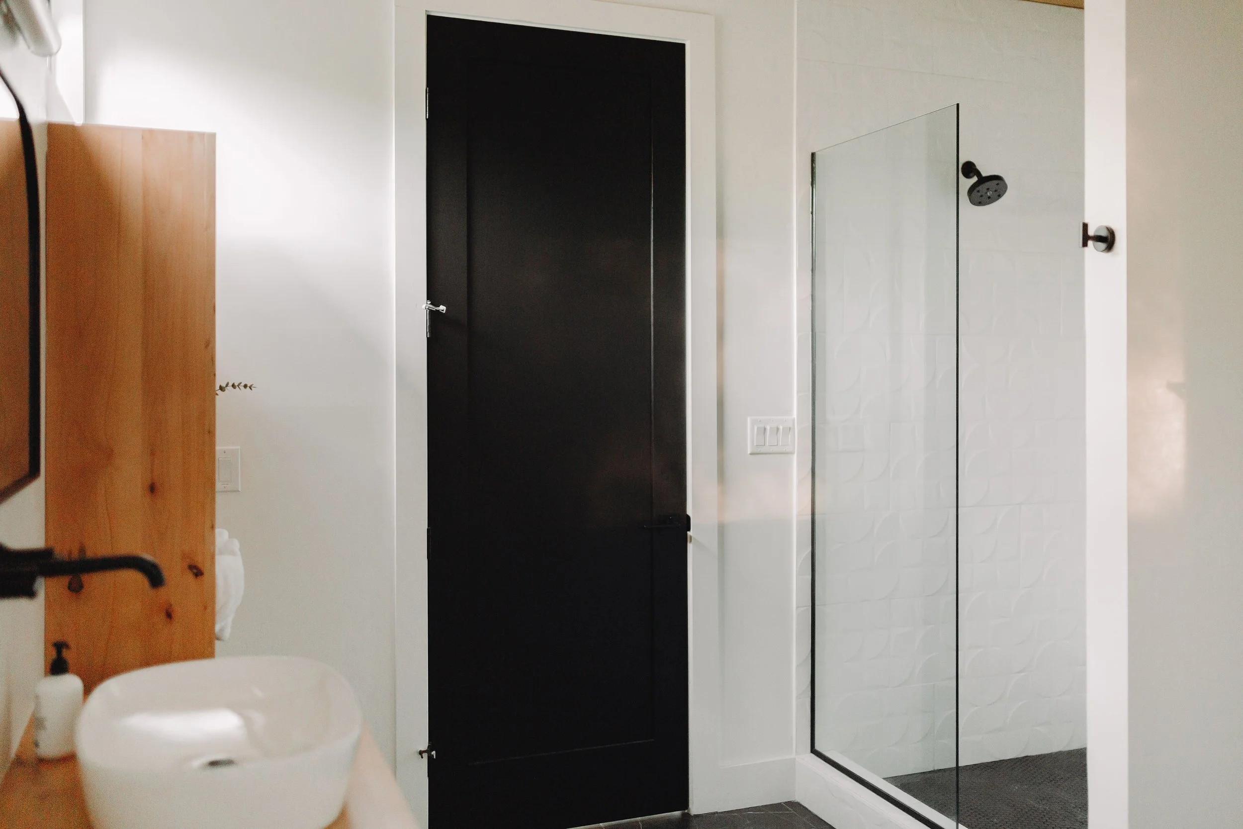 Modern bathroom with a black door, white sink, wooden shelf, and a glass walk-in shower with a black showerhead.
