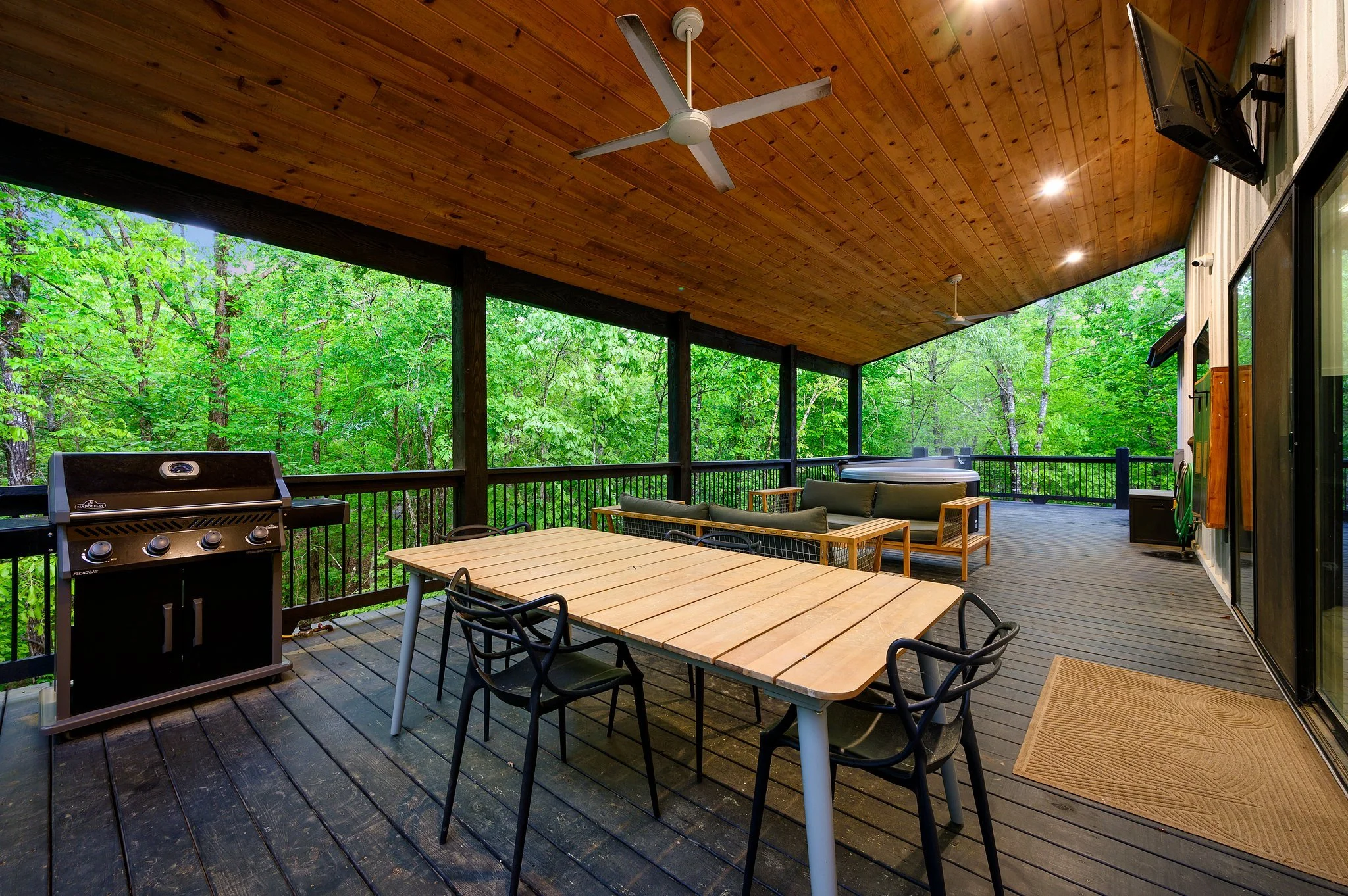 Screened outdoor deck with wooden ceiling, ceiling fan, black railing, table with four black chairs, lounge seating with cushions, hot tub, grill, and mountain view with lush green trees.