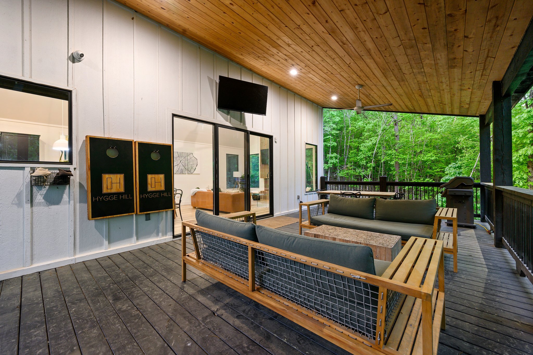 Outdoor covered patio with wooden ceiling, seating area with a sofa and chairs, a grill, and view of green trees.