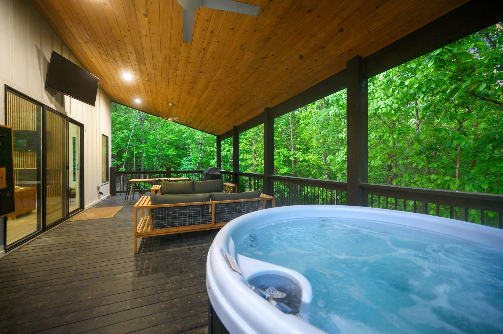 A covered outdoor balcony with a hot tub, seating area with couches, a table with chairs, surrounded by lush green trees.