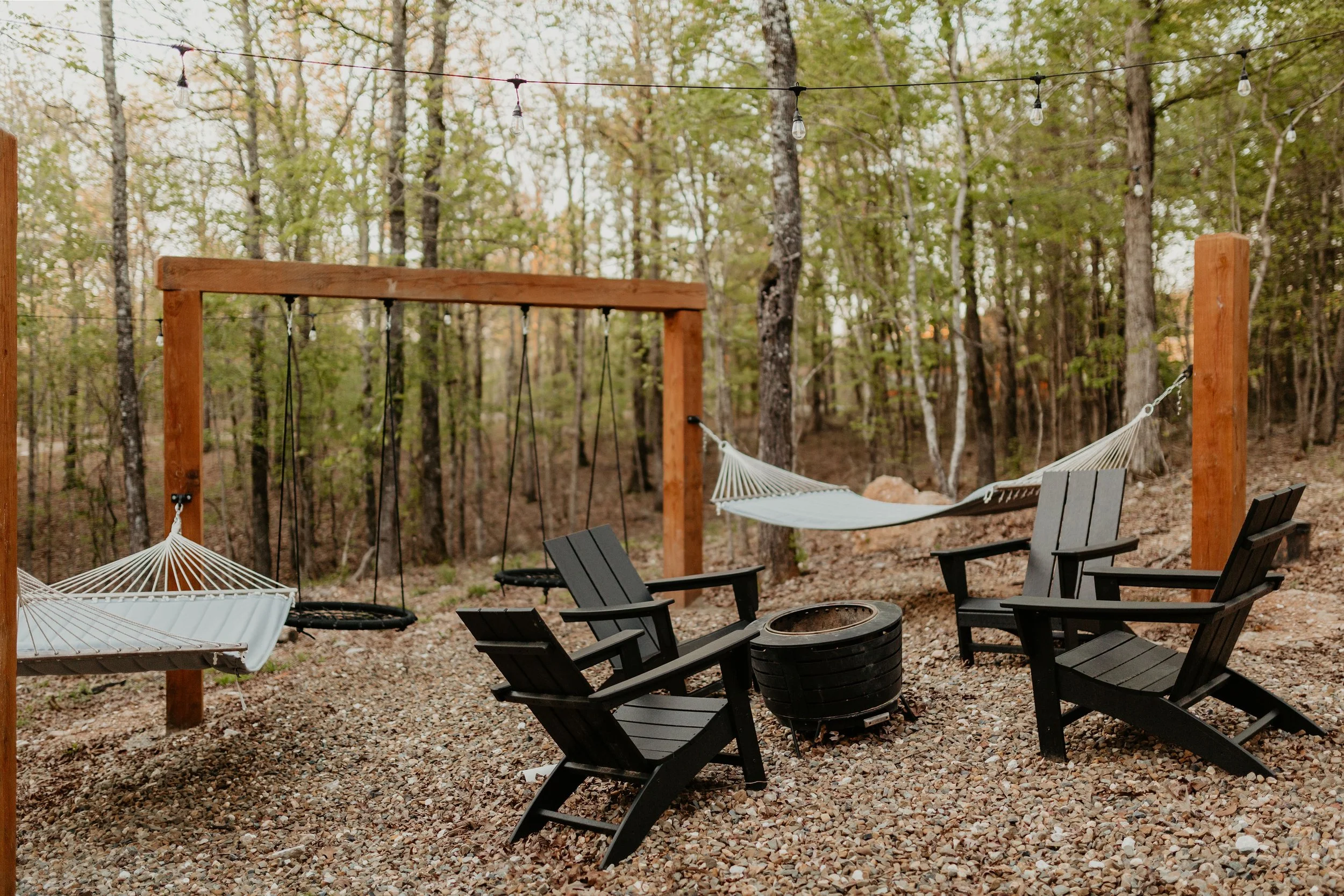 Outdoor backyard with gravel ground, wooden chairs, a fire pit, hammock, swings, and string lights, surrounded by trees.