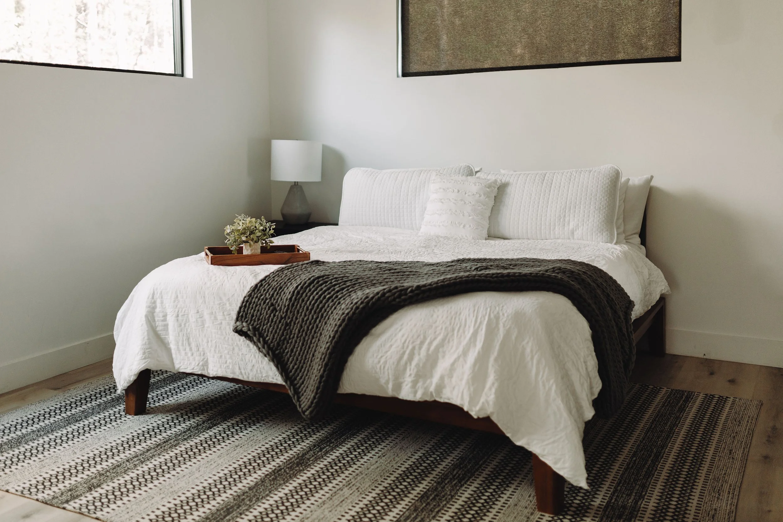 Modern bedroom with a white bed, black throw blanket, and a tray with a plant, a lamp on a side table, and two windows on the wall.