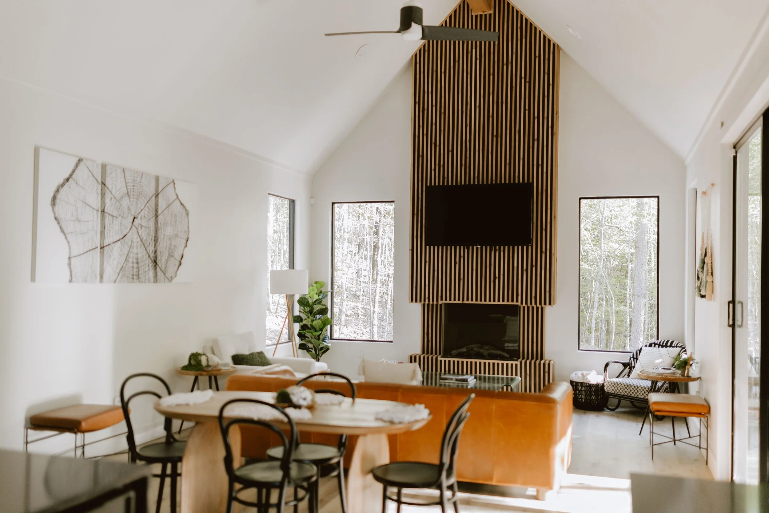 Modern living room in Hygge Hill, with a high vaulted ceiling, a wood-paneled fireplace and wall-mounted TV, large windows with a wooded view, and various seating including a white sofa, armchairs, and dining chairs around a wooden table.