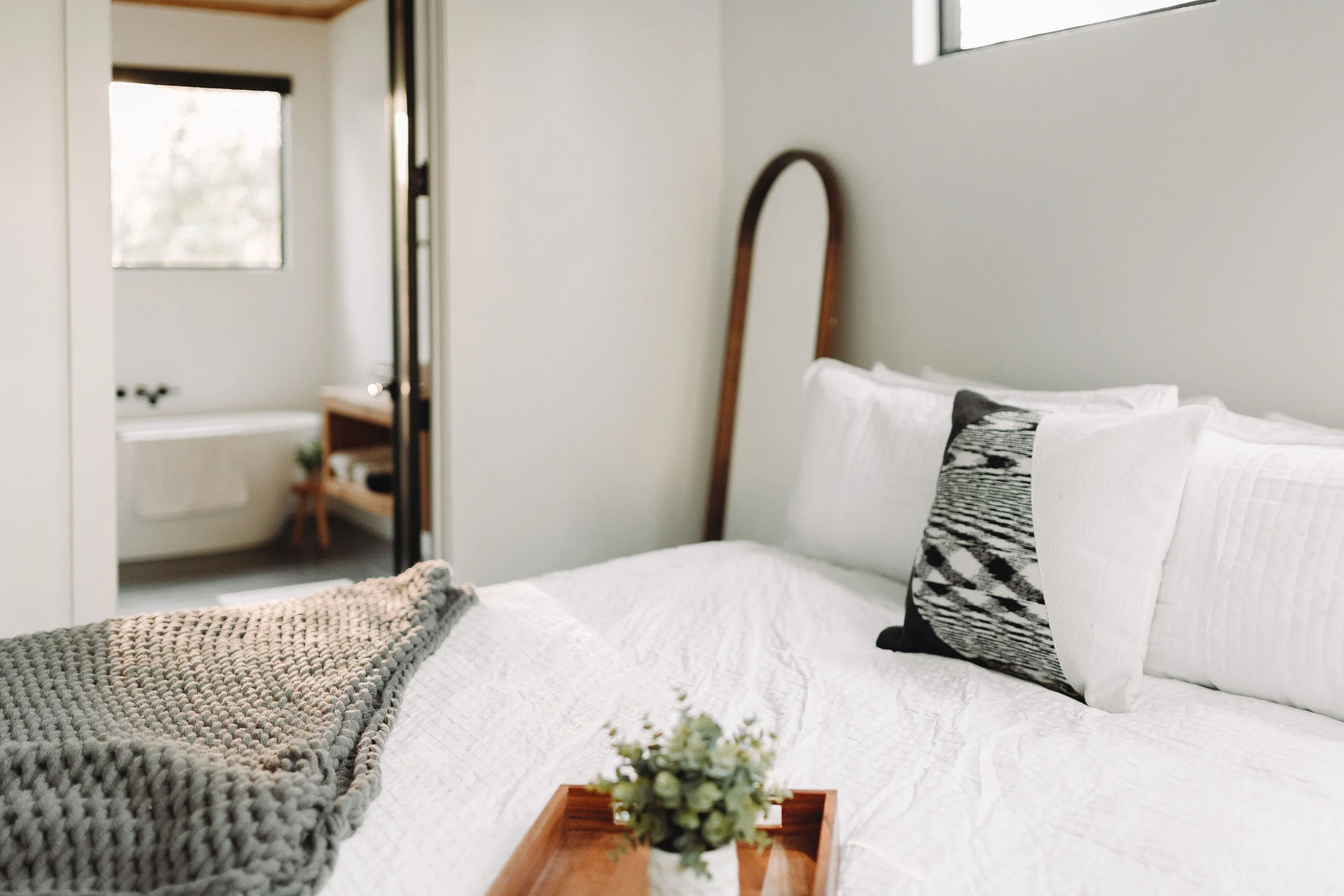 A cozy bedroom featuring a bed with white bedding, decorative pillows, a small tray with a plant, and a background view of a bathroom with a bathtub and a window.