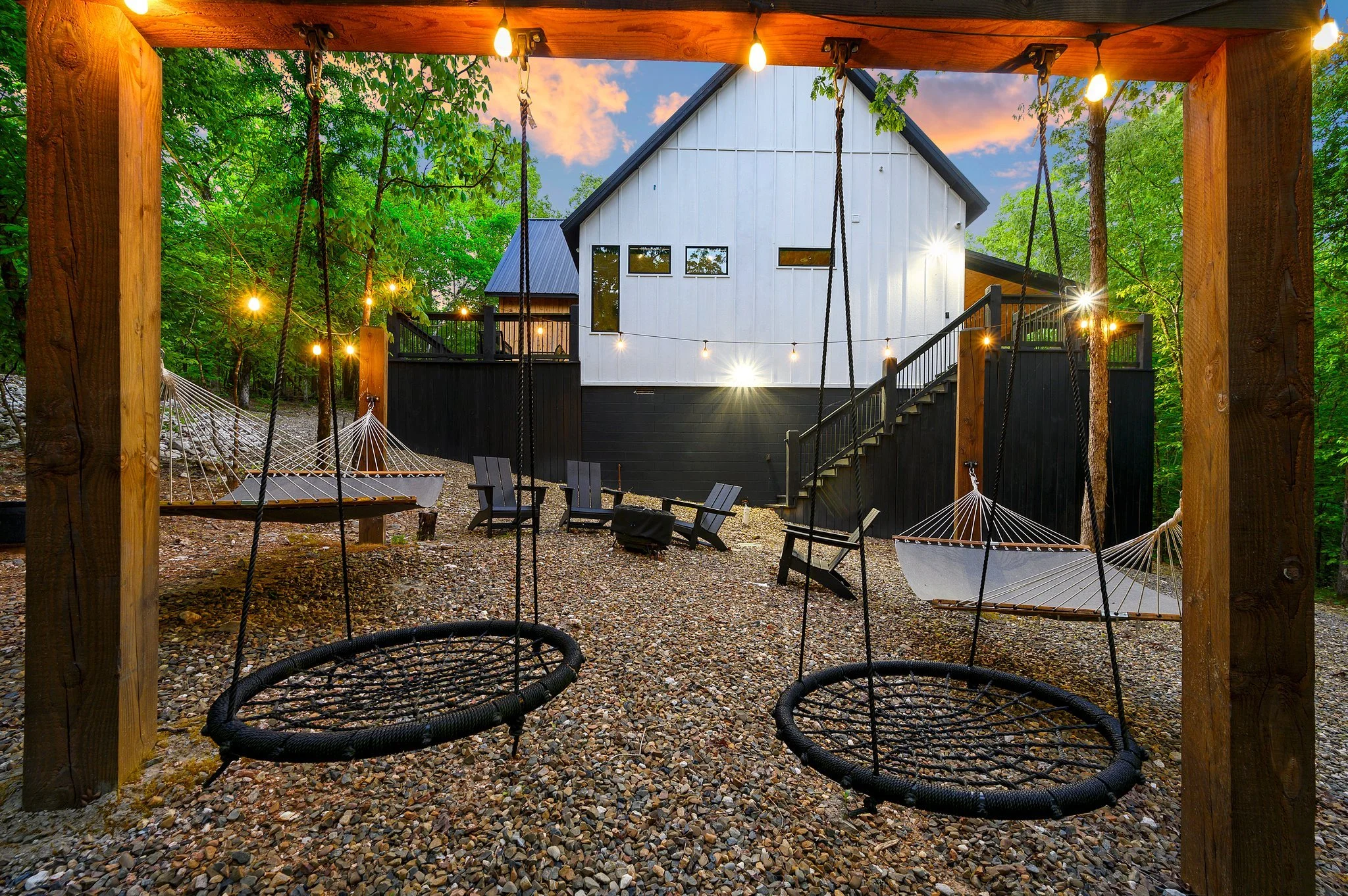Outdoor backyard at Hygge Hill, a modern cabin, with hanging round swings, black Adirondack chairs, string lights, surrounded by trees.