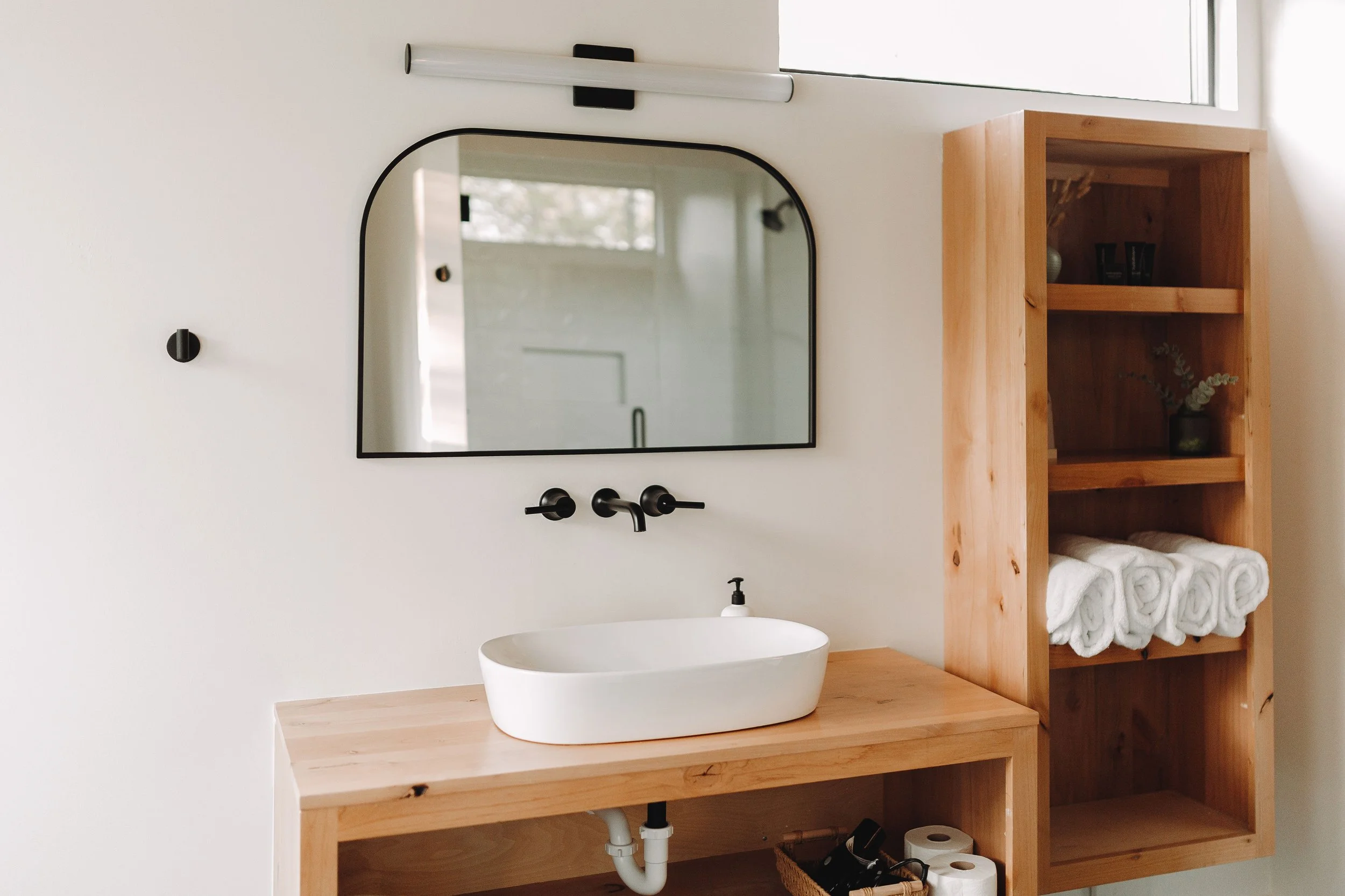 Minimalist bathroom with a wooden vanity, an oval vessel sink, black wall-mounted faucet, a black-rimmed mirror, and a wooden open shelf with rolled white towels and decorative items.