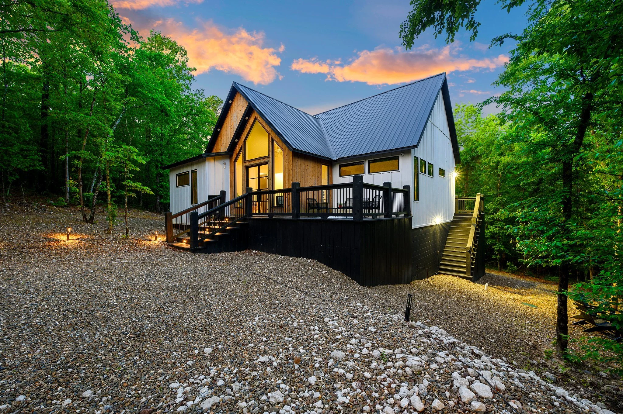 Hygge Hill, a modern cabin, with white and black exterior, large windows, and a metal roof, surrounded by lush green trees during sunset.