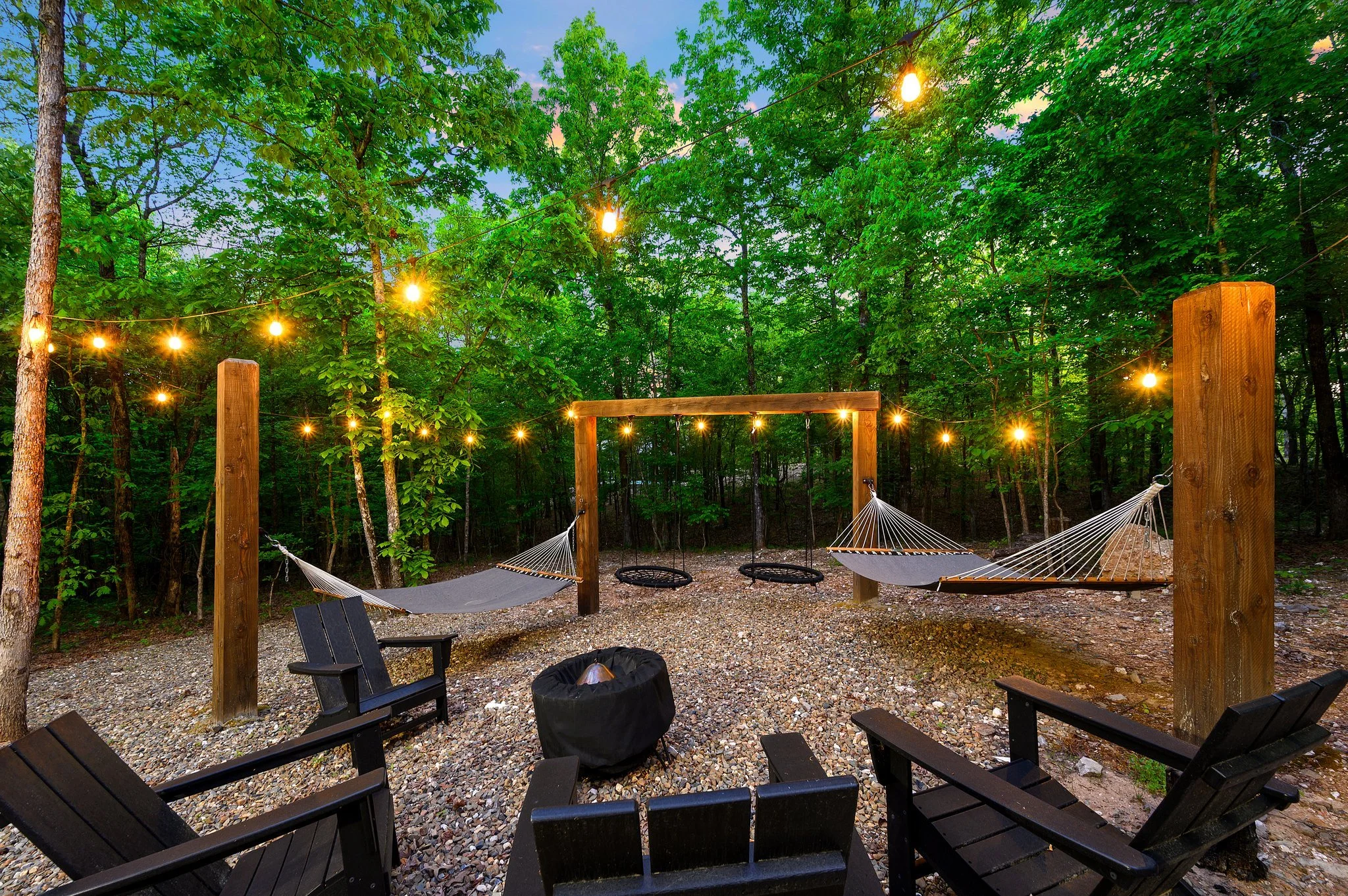 Outdoor backyard area with string lights, trees, and a gravel ground. There are two hammocks hanging between wooden posts, Adirondack chairs, and a fire pit. The scene is surrounded by dense green foliage.