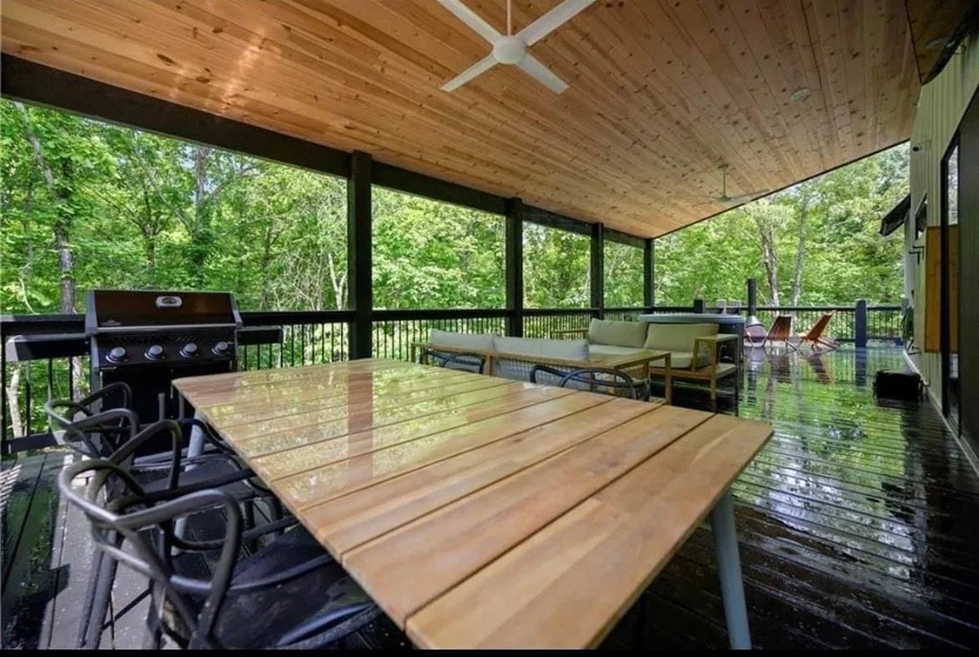 Covered outdoor porch with a wooden ceiling, black railing, and surrounded by green trees. Features a large wooden table with six chairs, a barbeque grill, a white sofa set, two orange lounge chairs, and a small black speaker.
